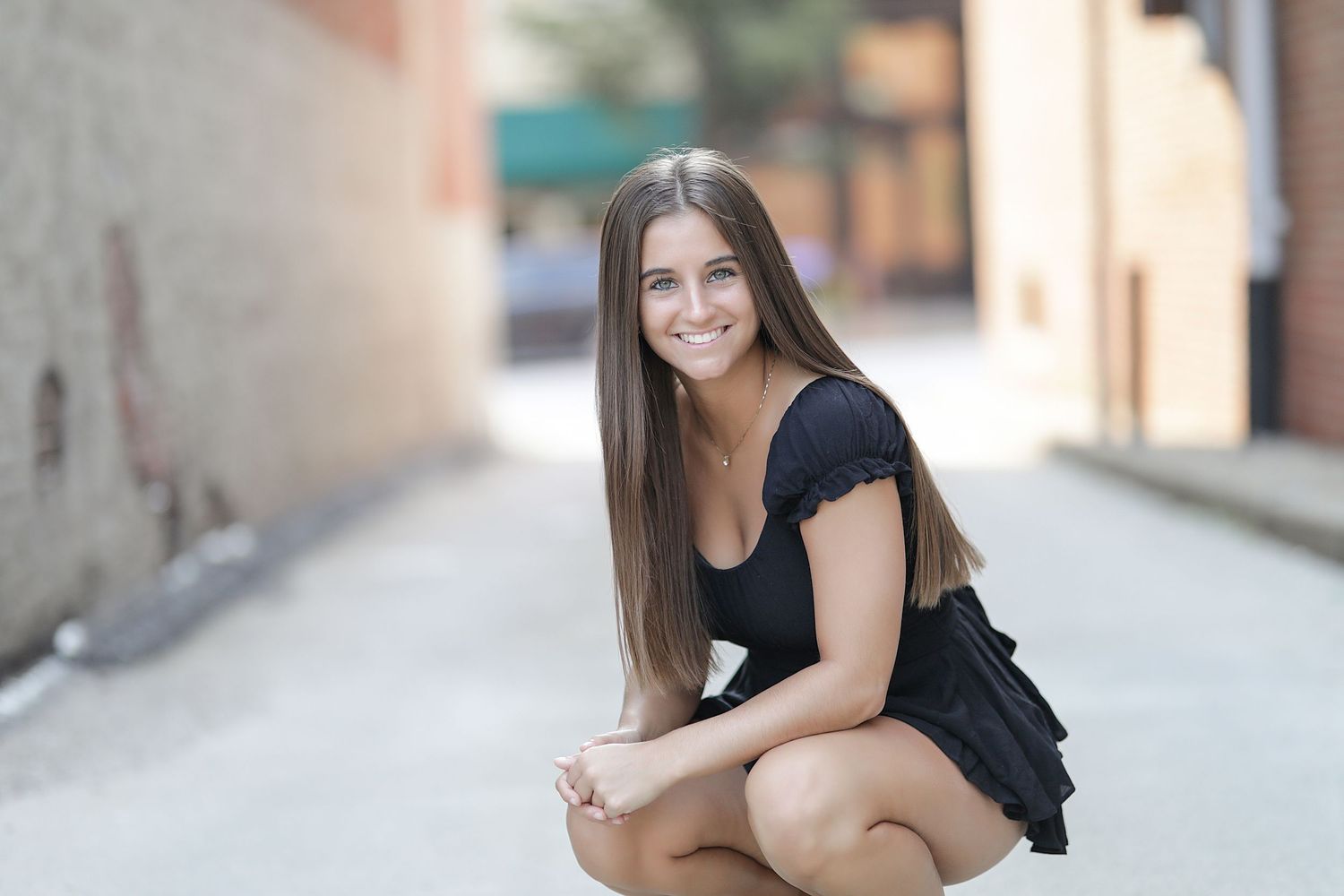 Outdoor portrait of someone in a black dress crouching in an urban alleyway with soft bokeh effect in background.