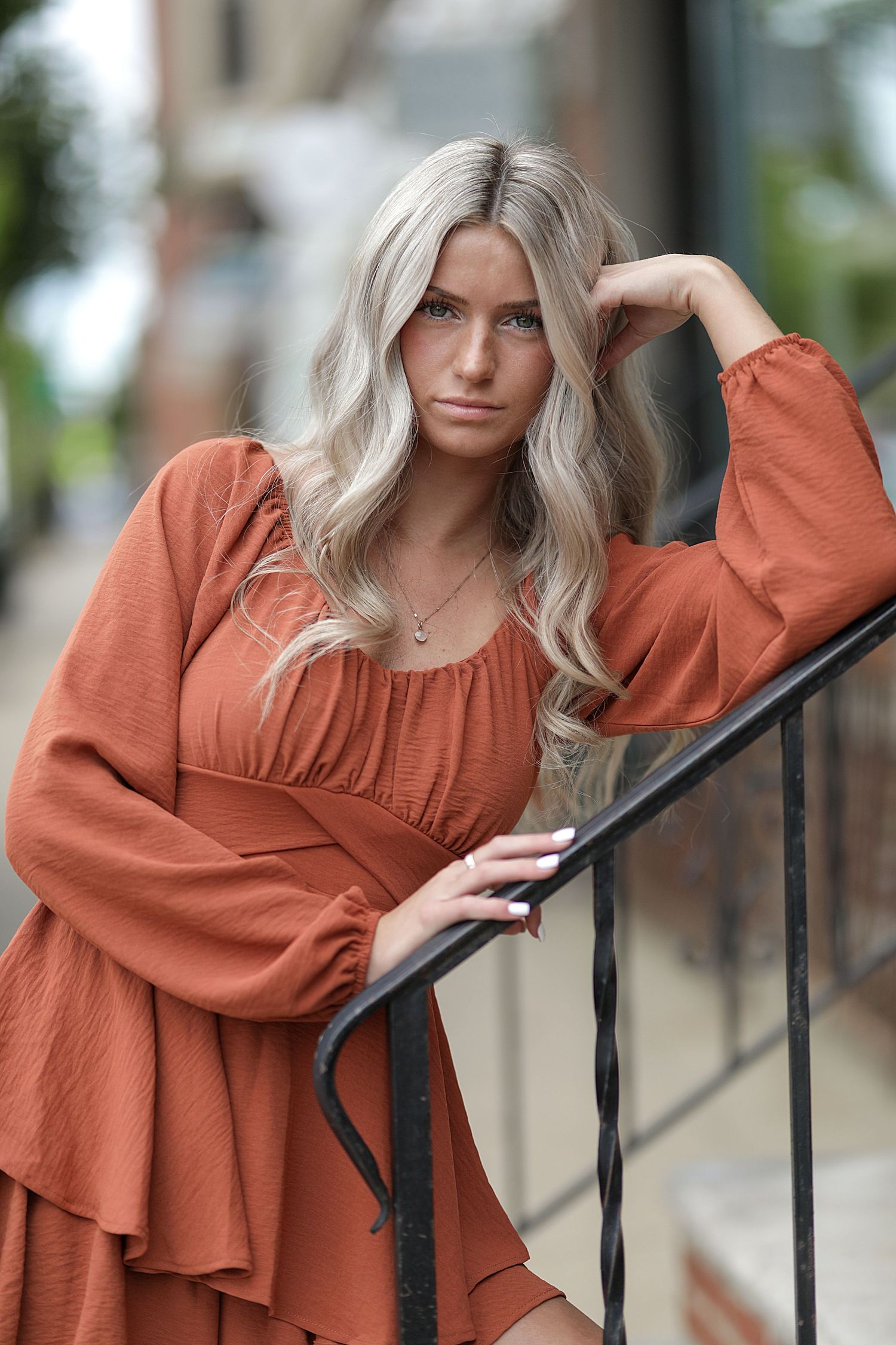 A person in a rust-colored flowy dress with balloon sleeves poses on outdoor stairs while touching their platinum blonde hair.