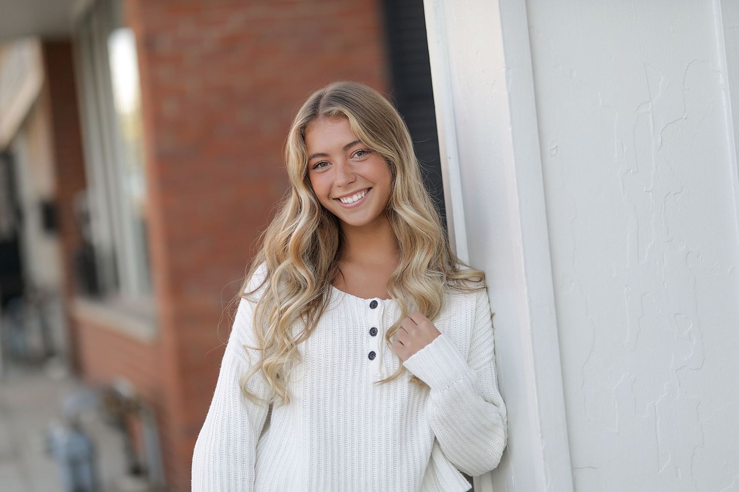 A woman wearing a cozy white knit sweater stands beside a brick wall while smiling outdoors.