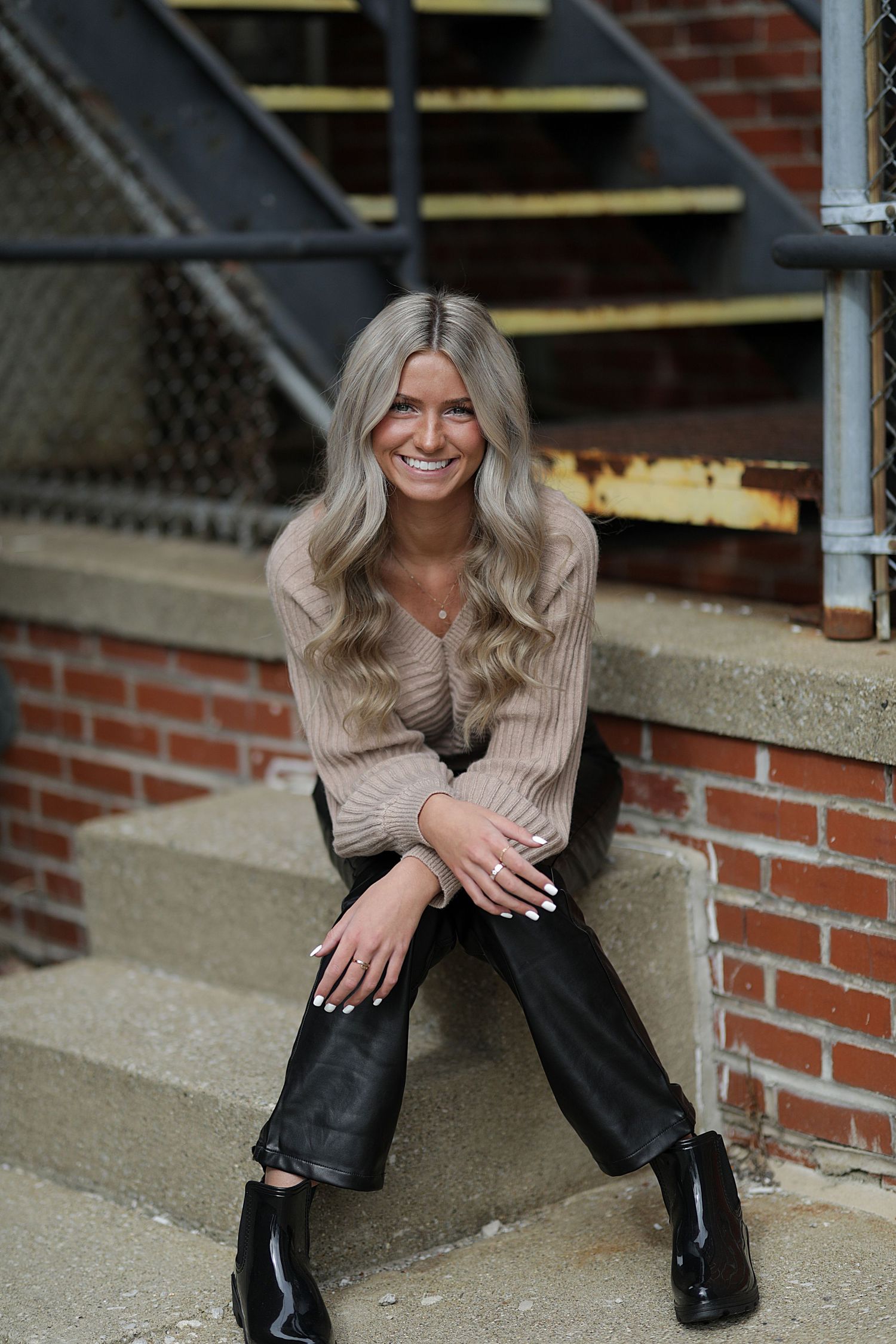 Someone in a beige knit sweater and black boots sits on brick steps with a natural smile.