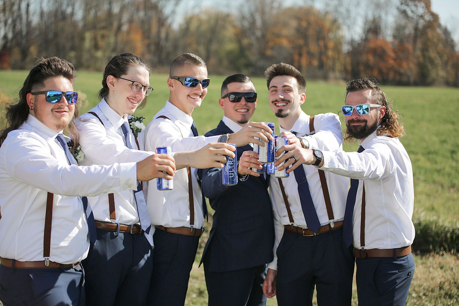 Groomsmen in white shirts and suspenders toast with drinks in an outdoor setting.
