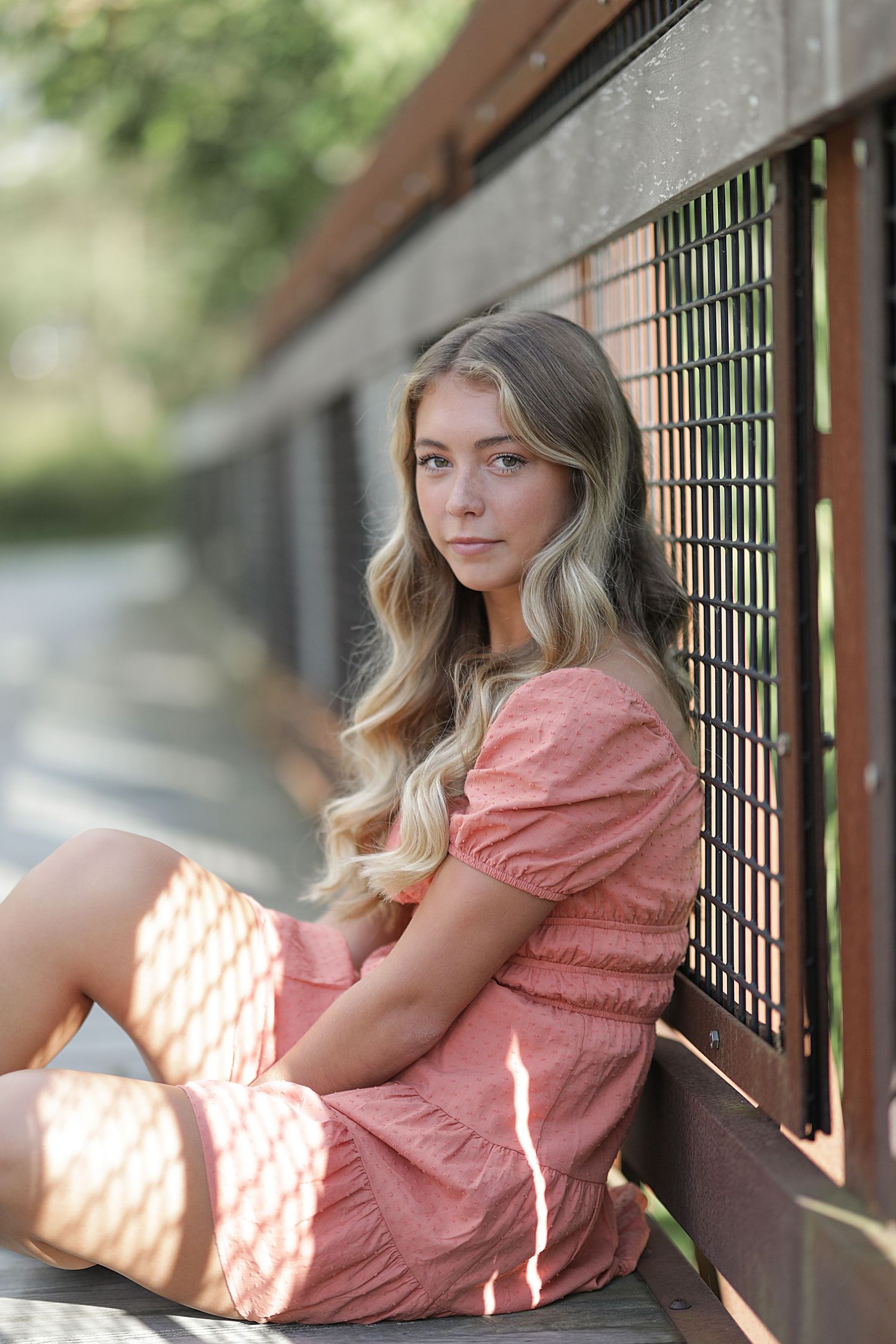 Young woman in coral pink dress sitting on wooden deck railing in dappled sunlight.
