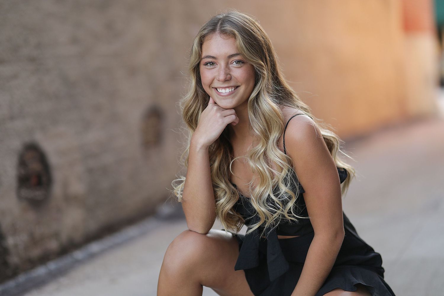 Young woman in black dress sitting outdoors with warm smile and long wavy blonde hair.