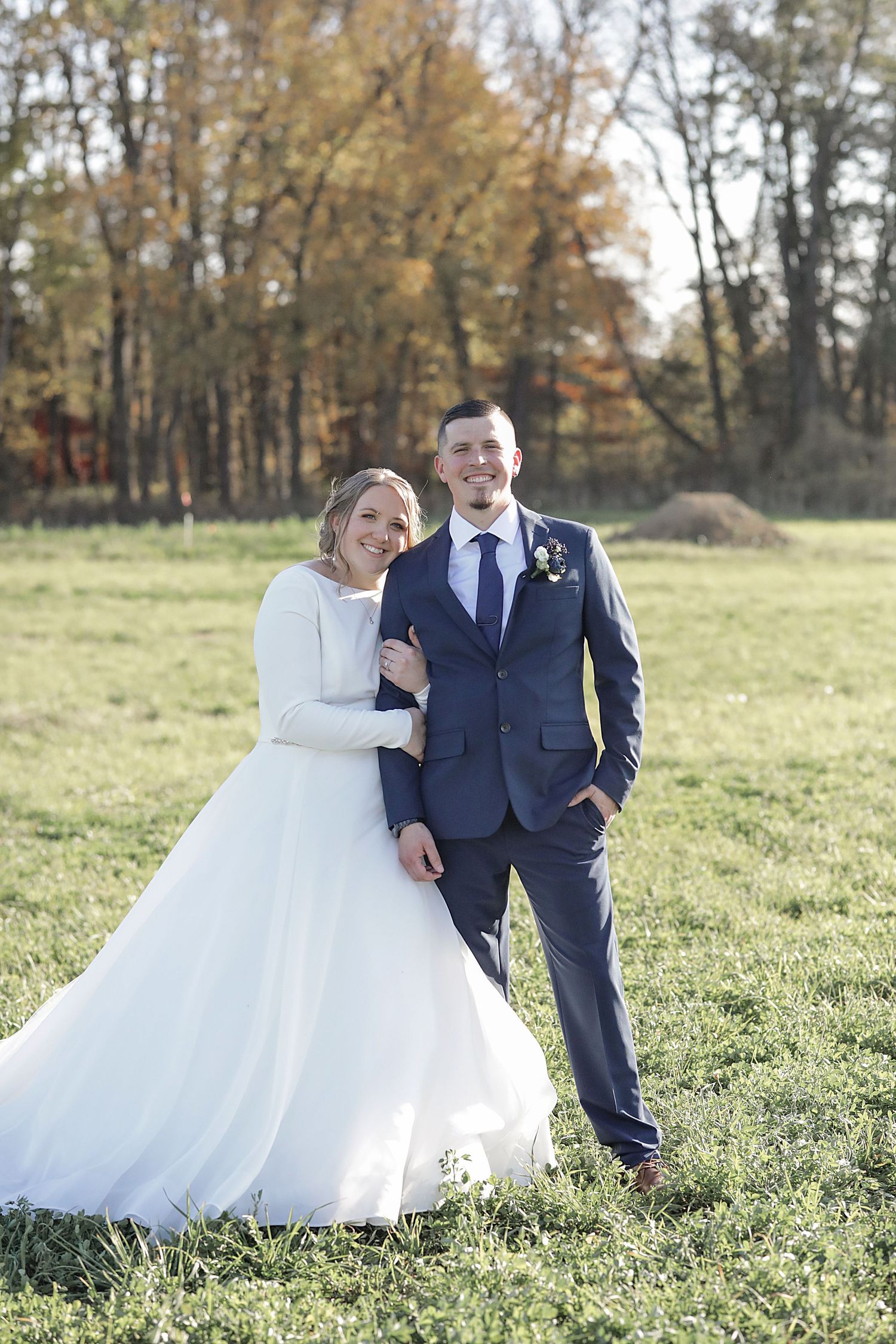Newlyweds pose together in an autumn field with golden trees in background.