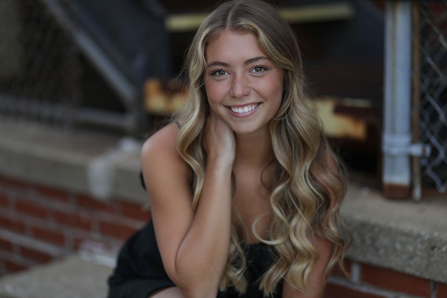 Professional outdoor portrait of a woman with long wavy blonde hair wearing a black dress against a brick wall background.