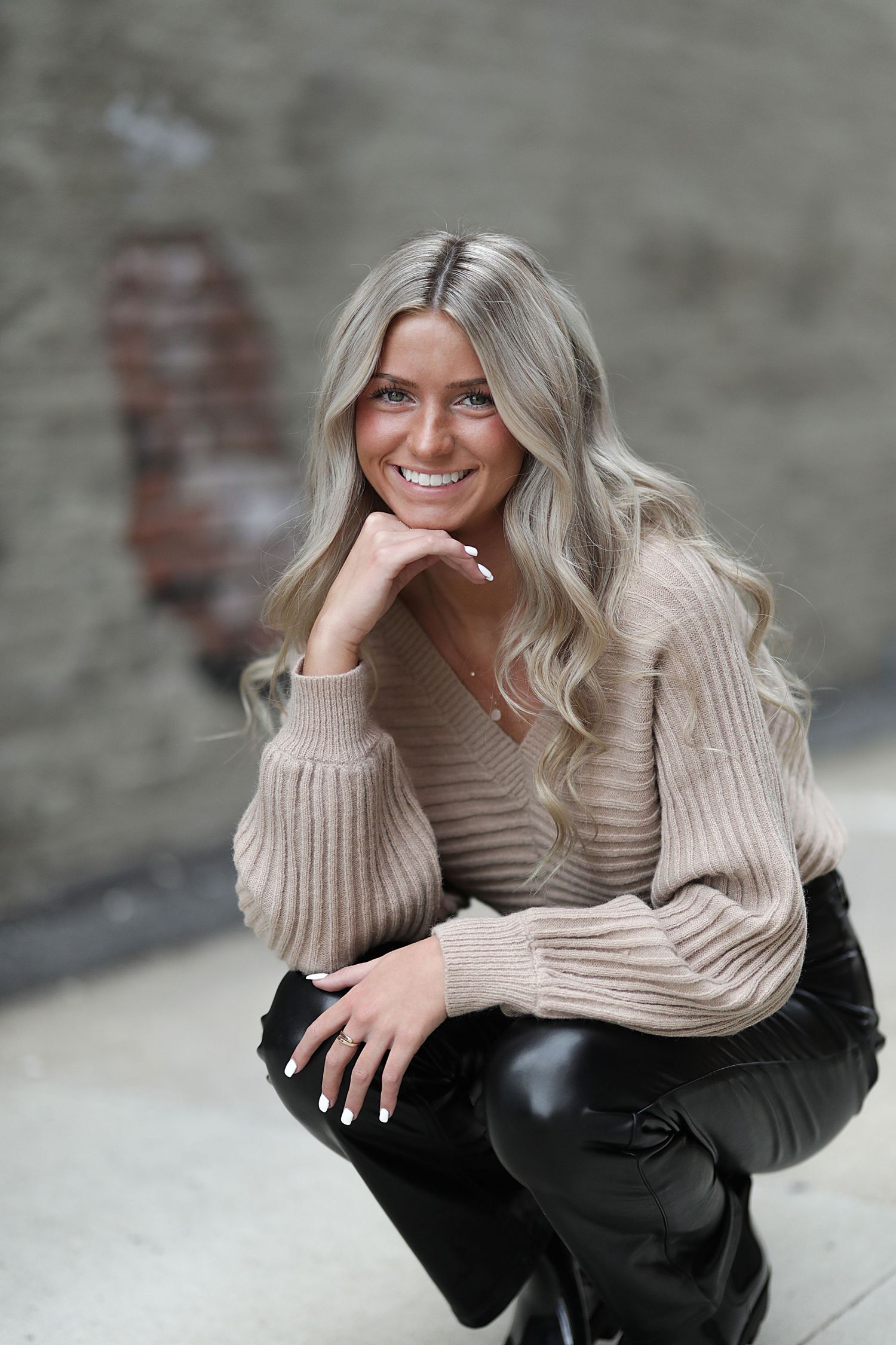 Stylish portrait of a person in a beige sweater and black pants crouching with a warm smile against a gray wall.