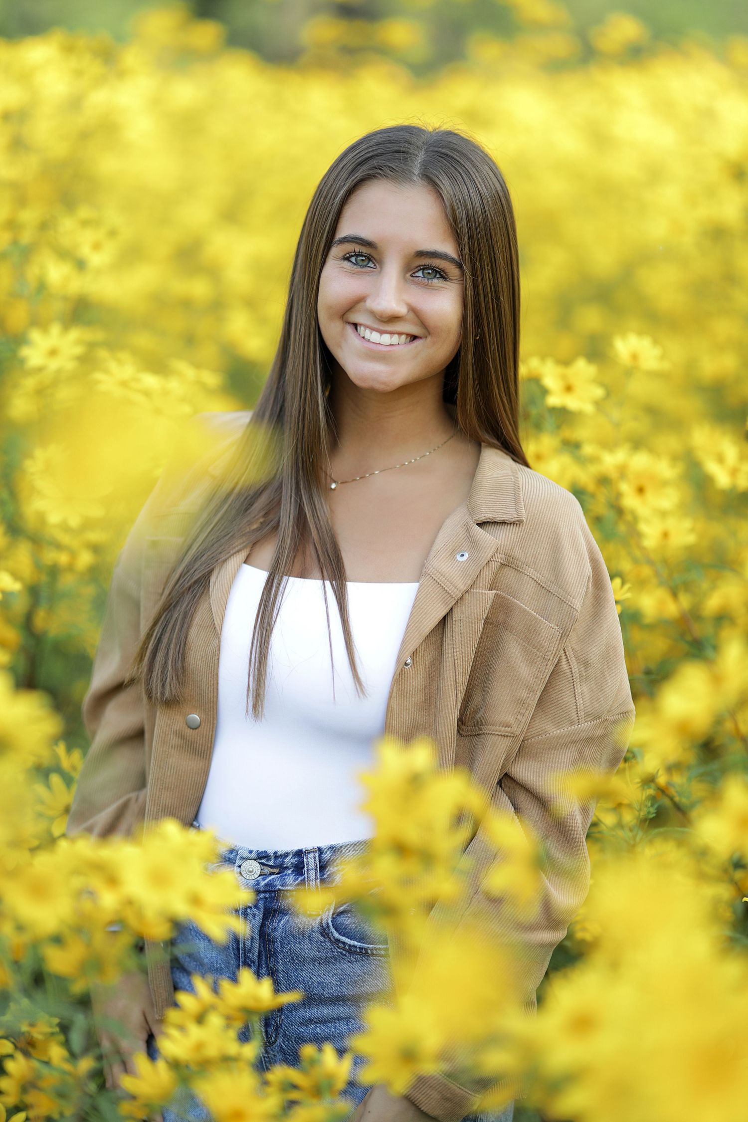 Portrait in a field of vibrant yellow wildflowers wearing a tan jacket and white top against a sunny natural background.