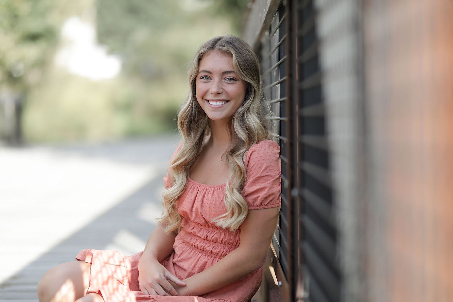 Someone in a pink dress with wavy blonde hair sits on a porch and smiles while leaning against a wall.