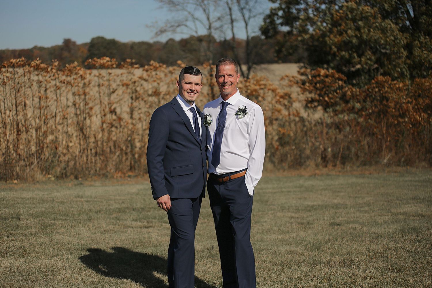 Two people in formal attire stand together in a field during autumn.