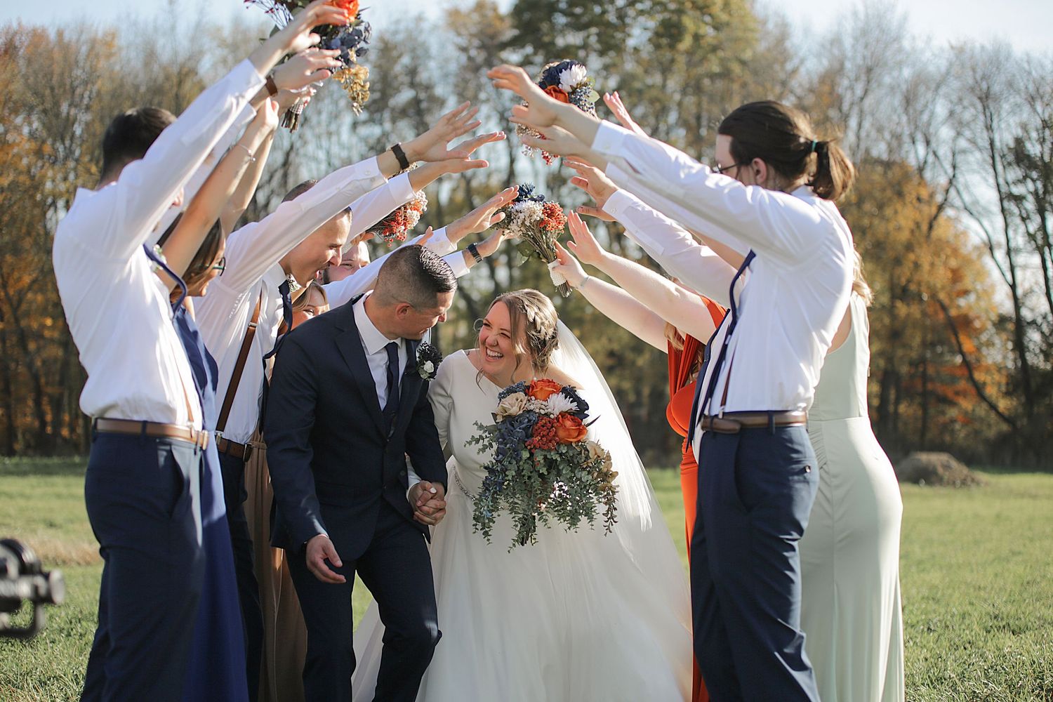 Wedding party creates an arch with raised arms for newlyweds to walk through in autumn setting.