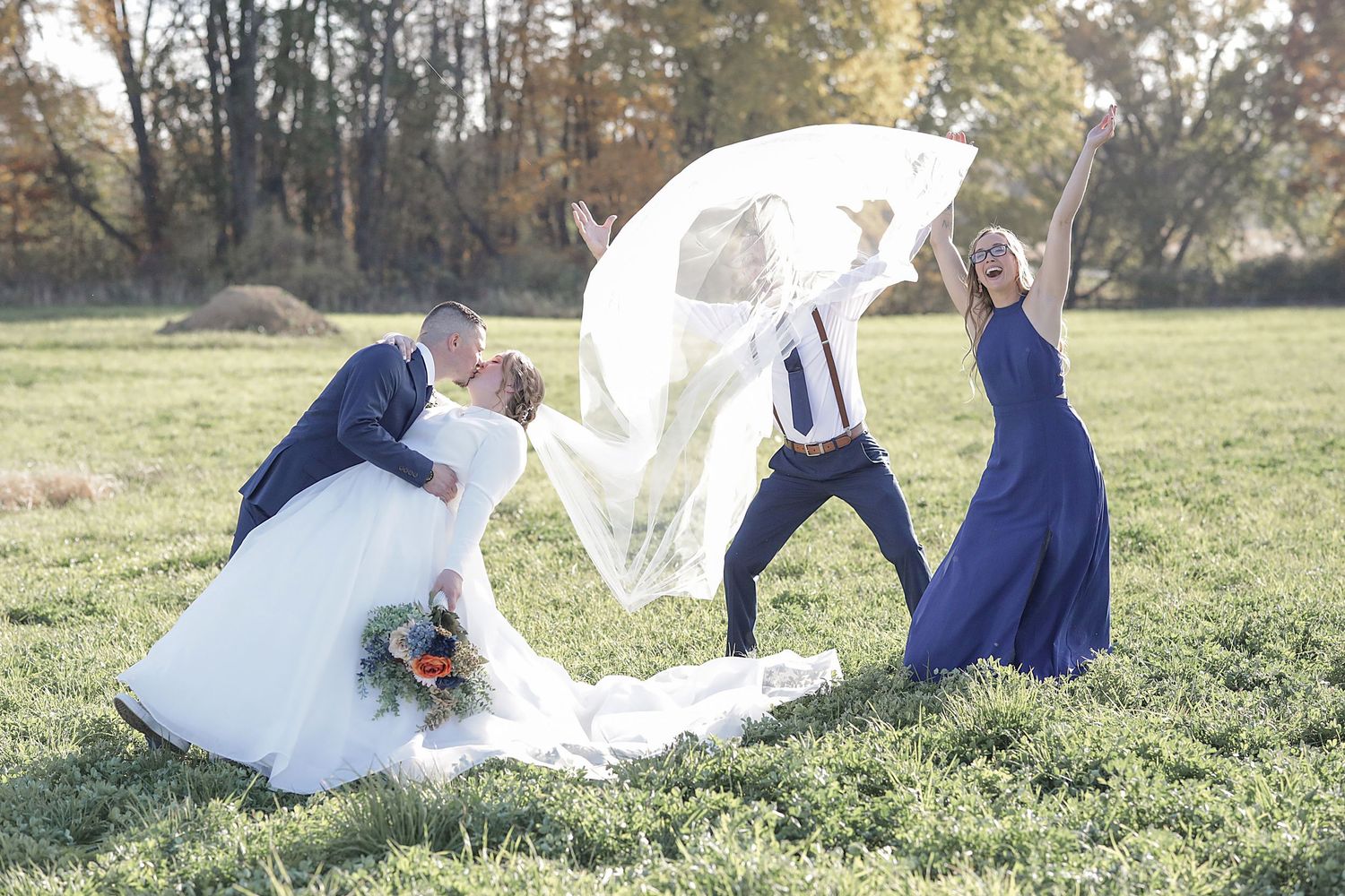 Wedding party members playfully catch a flowing veil in the wind on a grassy field.