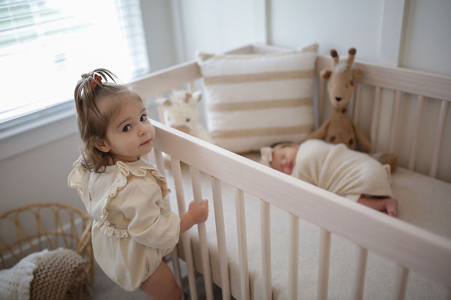Baby nursery decorated in neutral tones with vintage-style crib and white curtains beside a large window.