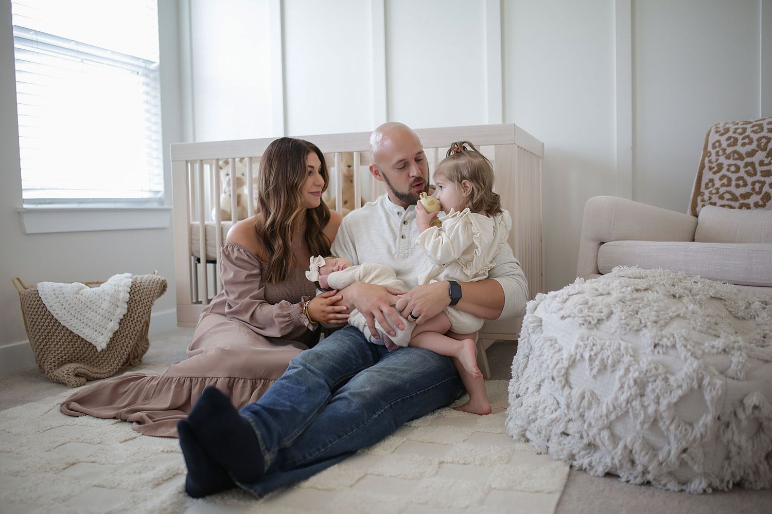 A family sits together on the floor of a cozy white bedroom with textured bedding and natural light from a window.
