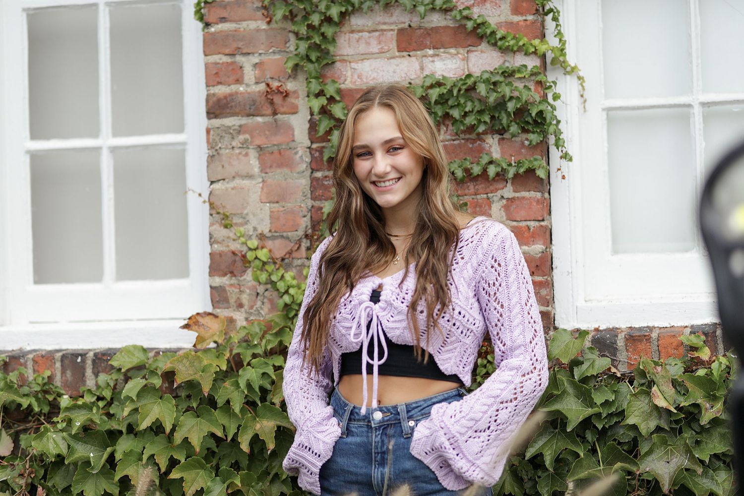 Casual fashion pose against rustic brick wall with climbing vines and white windows.