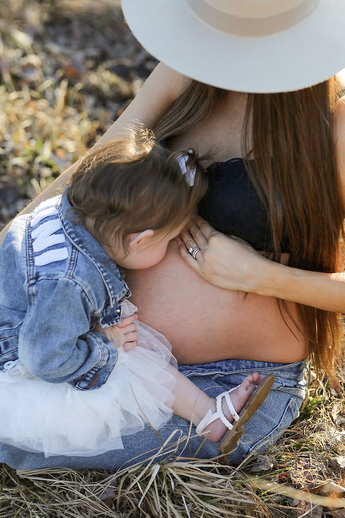 A sweet maternal moment captured outdoors as a child in denim leans against a pregnant belly in natural light.
