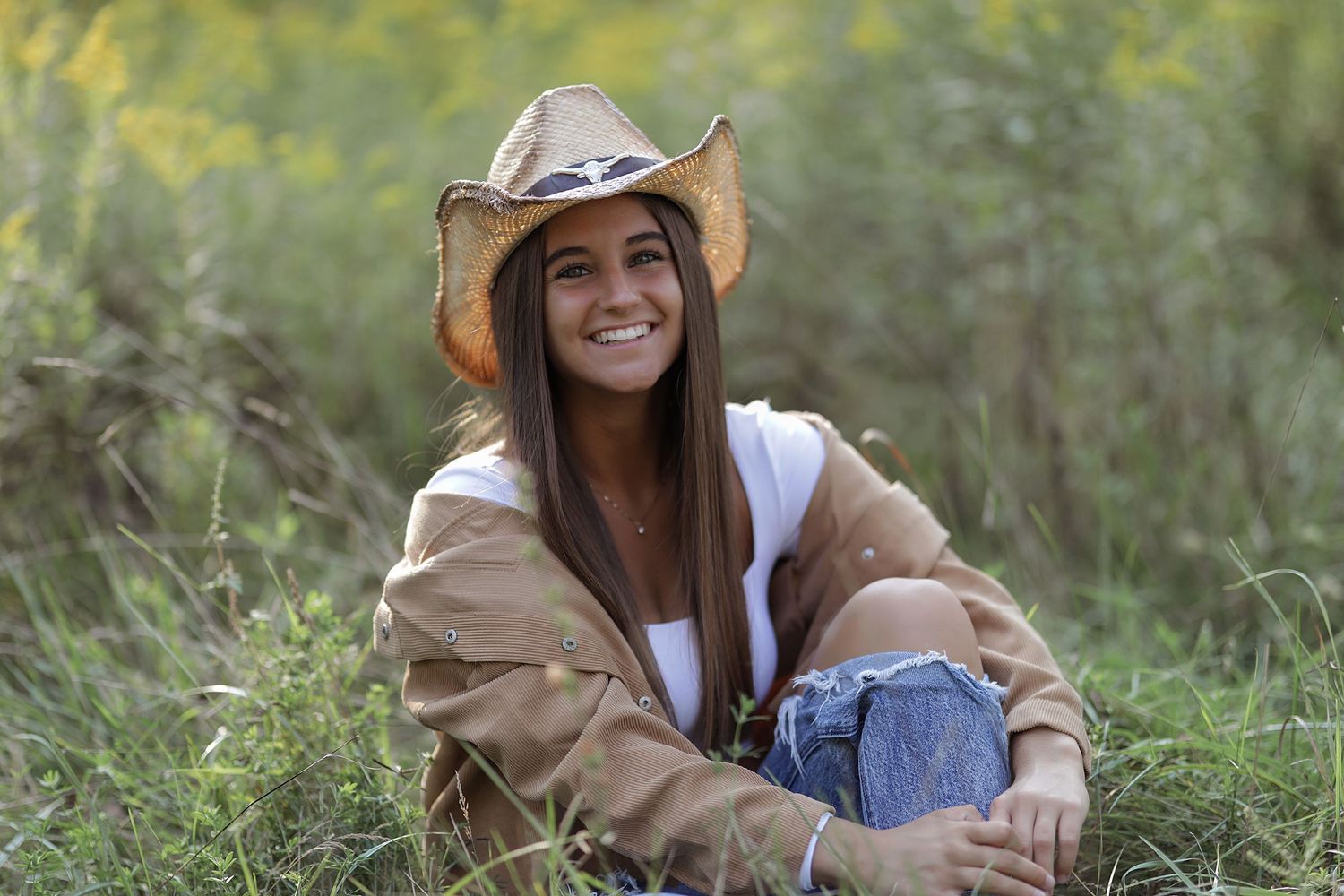 A country style portrait in a grassy field featuring someone wearing a tan jacket, cowboy hat and blue jeans.