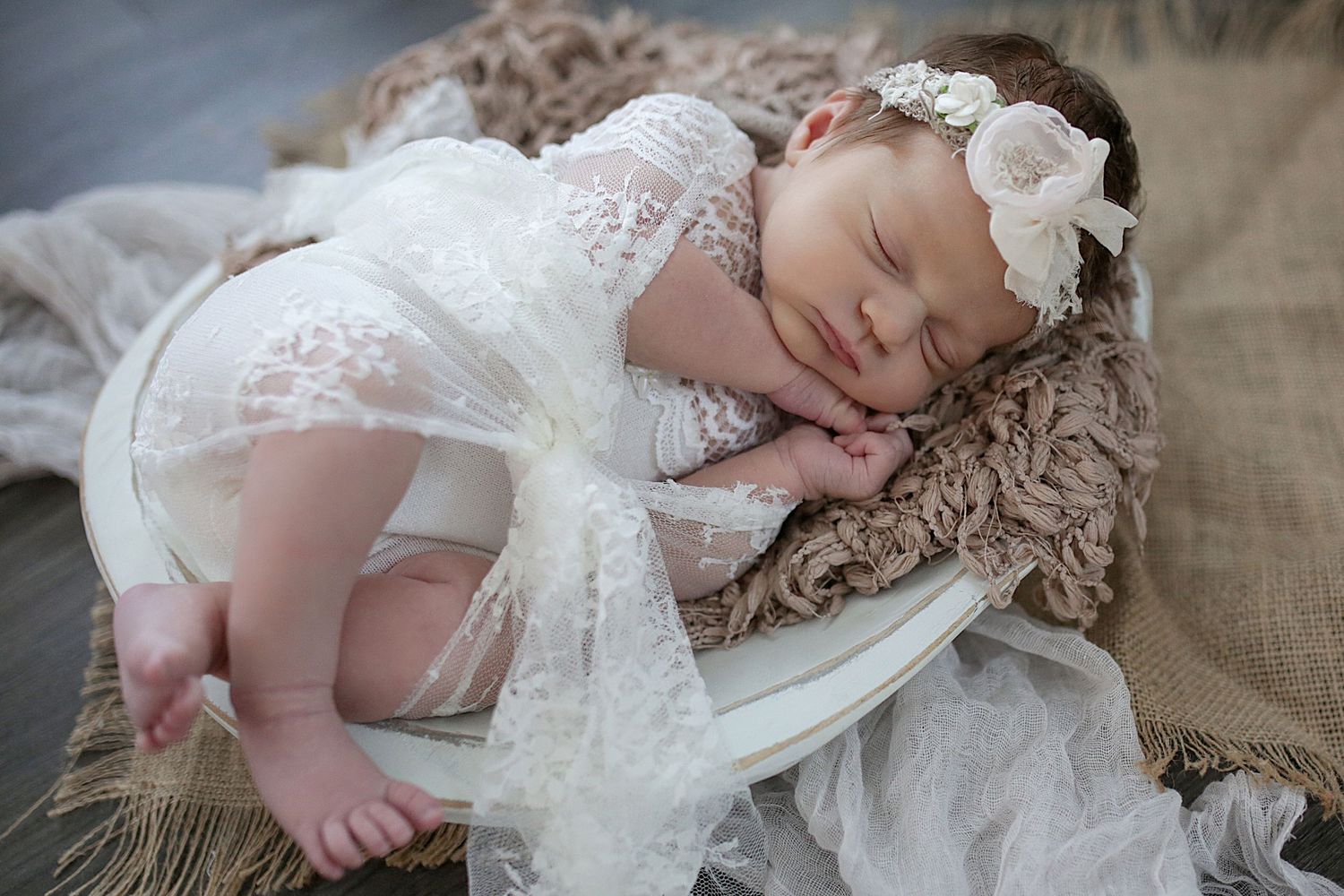 A sleeping baby wearing a white lace dress and floral headband rests peacefully on a vintage-style chair.