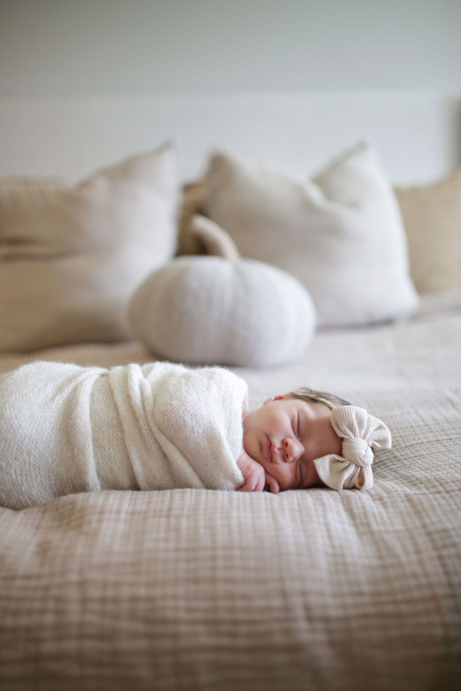 A sleeping newborn wrapped in soft white fabric lies peacefully on a neutral-toned bed with white pillows in the background.
