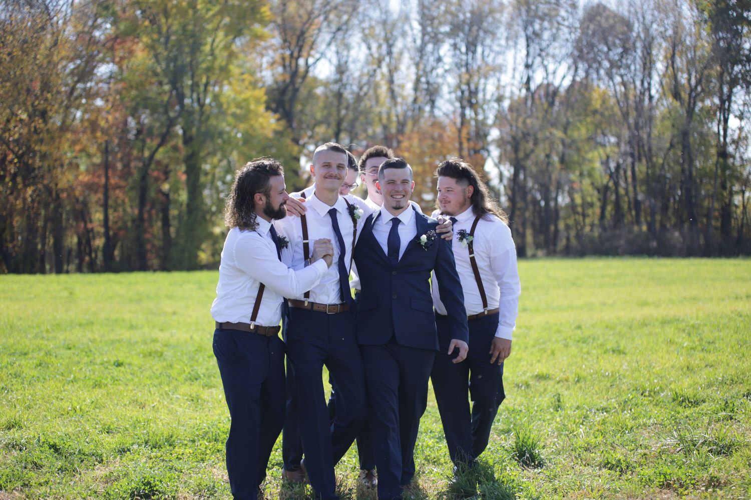 Group of groomsmen in suspenders pose together in an autumn field.