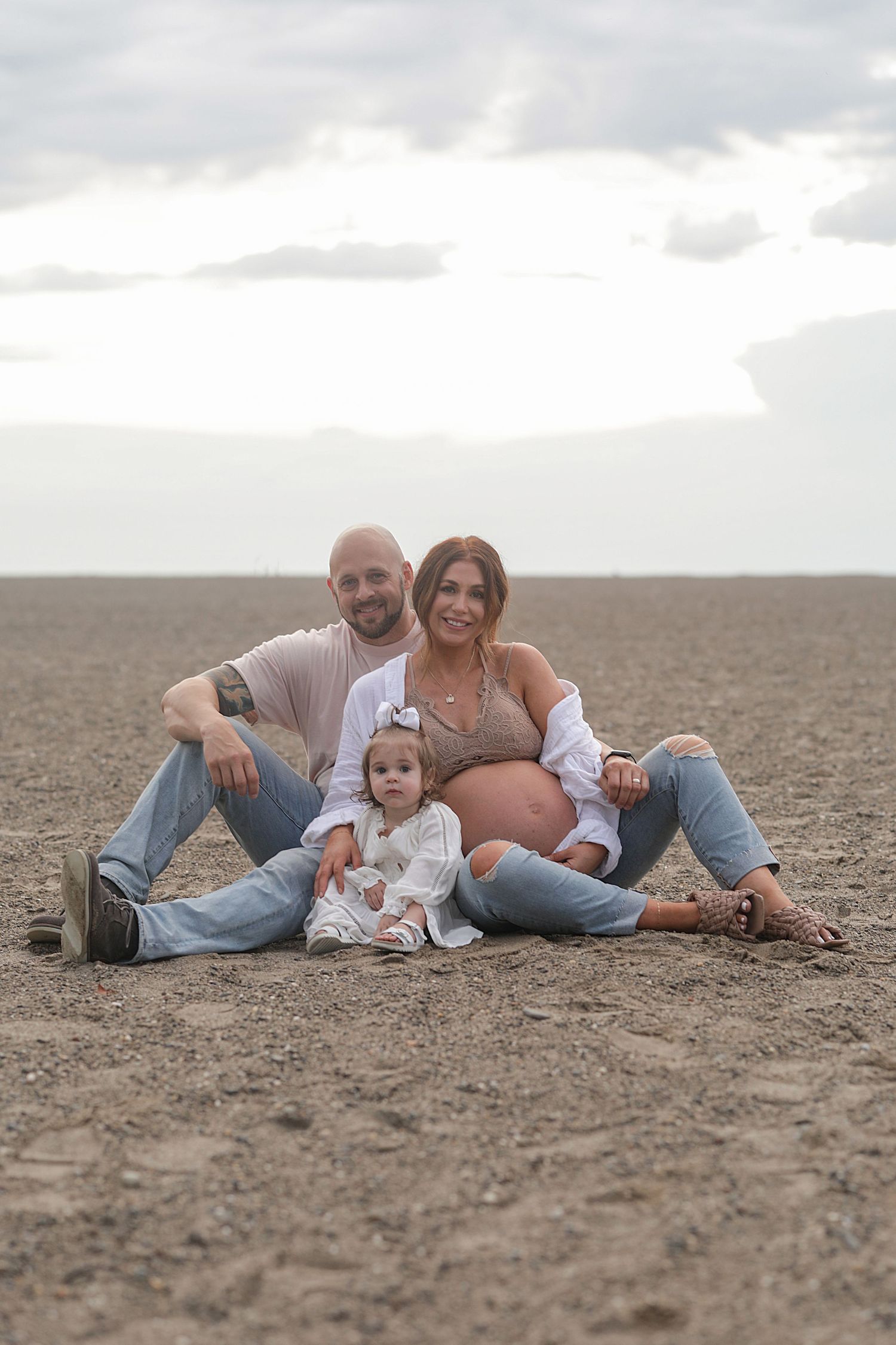 A family sits together on sandy ground wearing casual denim and white clothing against a cloudy sky.