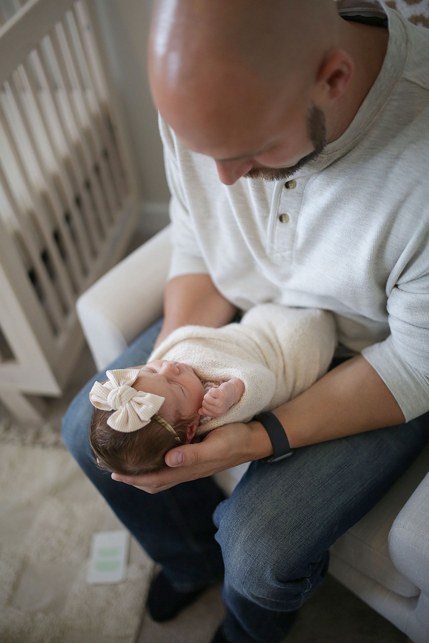 A parent holds a sleeping baby swaddled in a white blanket while sitting on a couch in soft natural light.