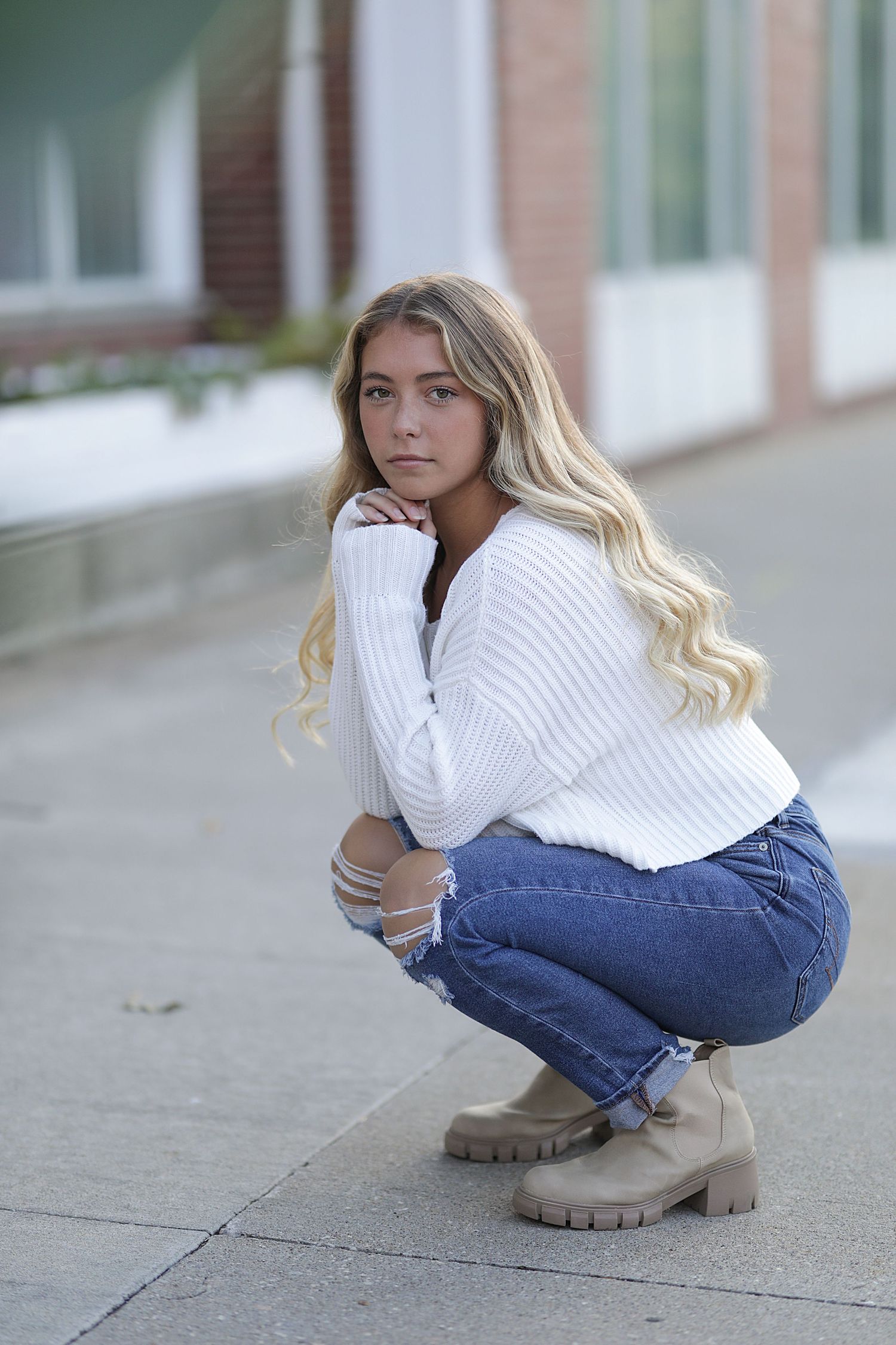 A woman in a white knit sweater and jeans crouches on the sidewalk wearing chunky beige boots.