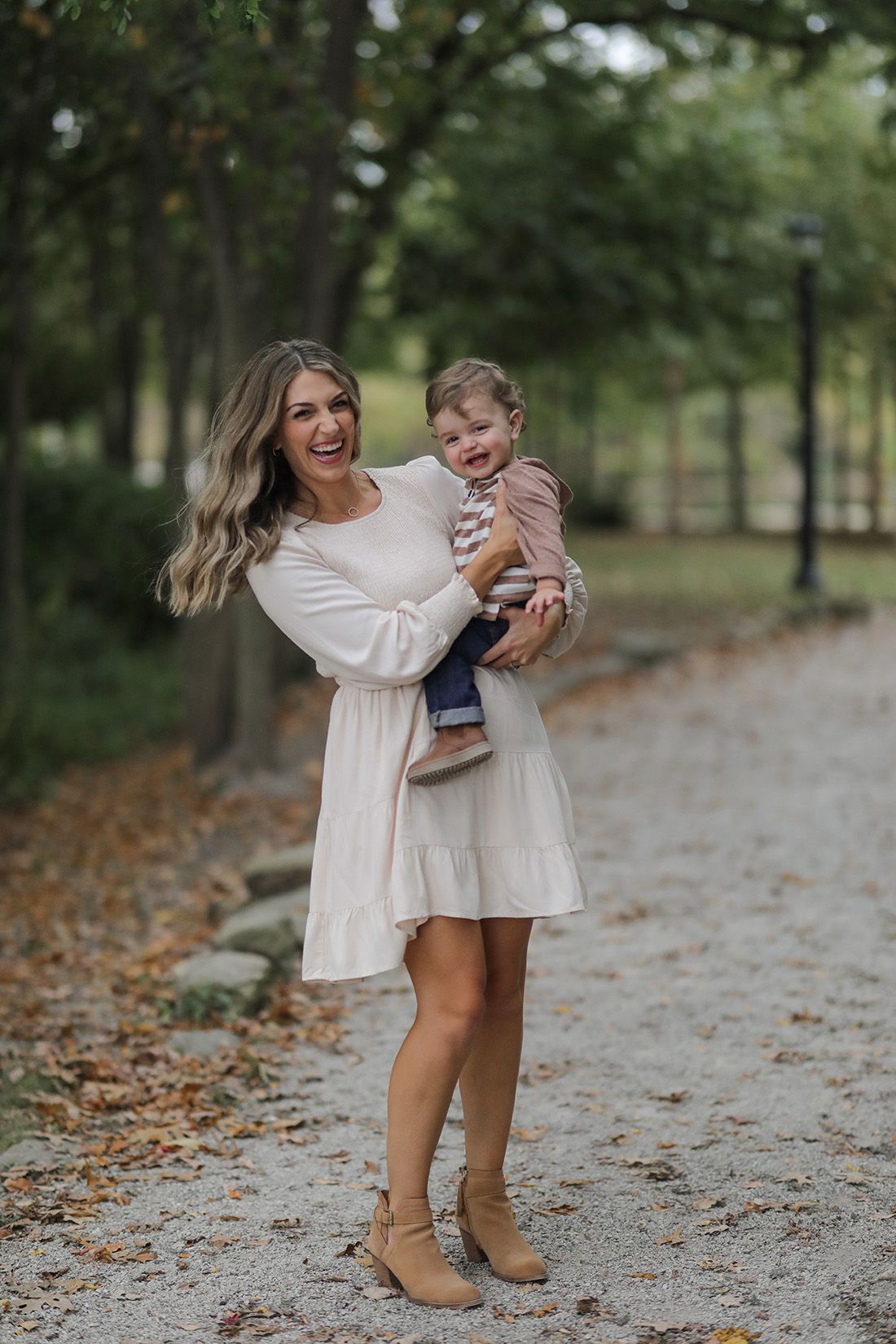 Joyful parent in white dress twirls with toddler on autumn path surrounded by trees.