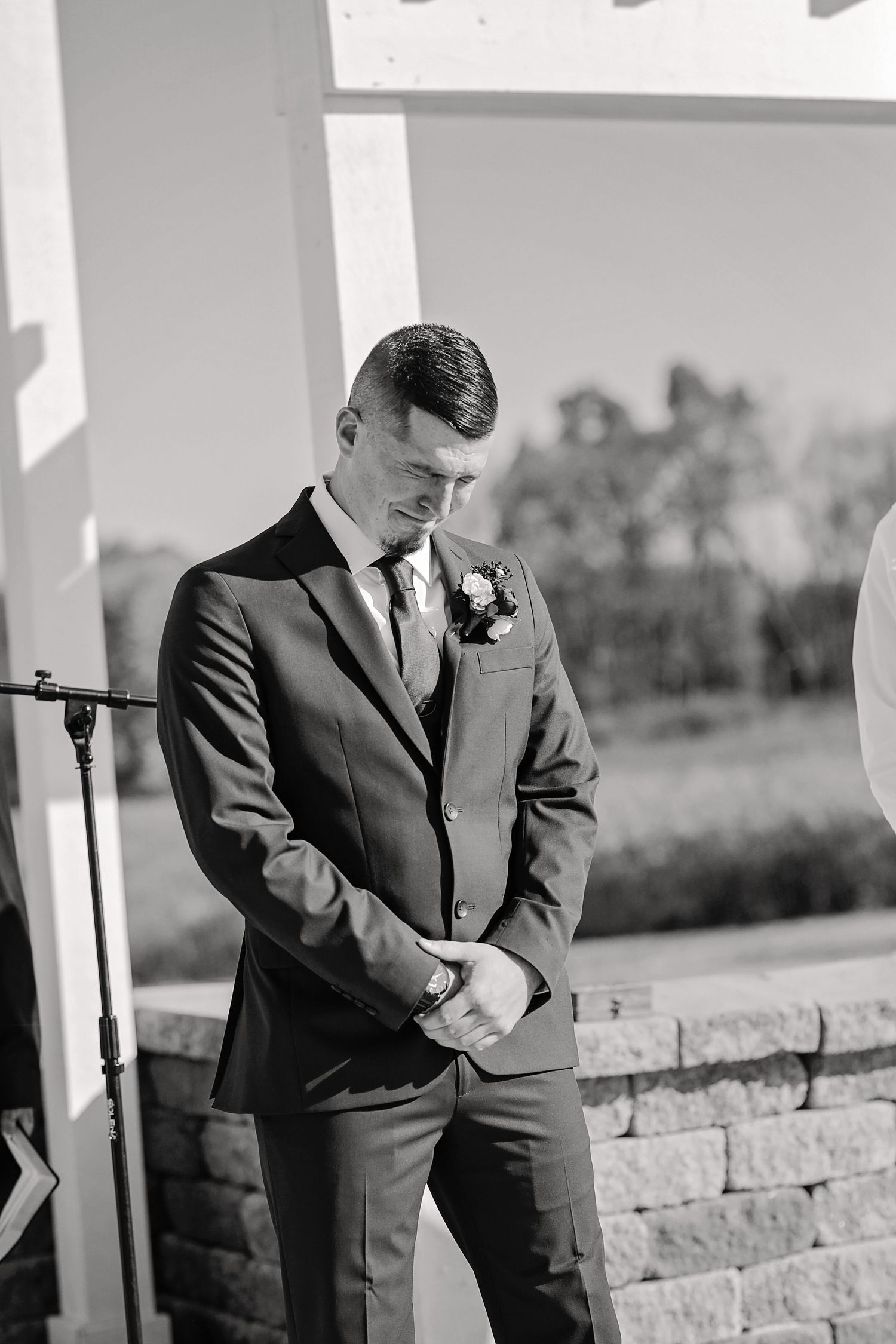 A groom in a fitted dark suit with boutonniere stands contemplatively in black and white.
