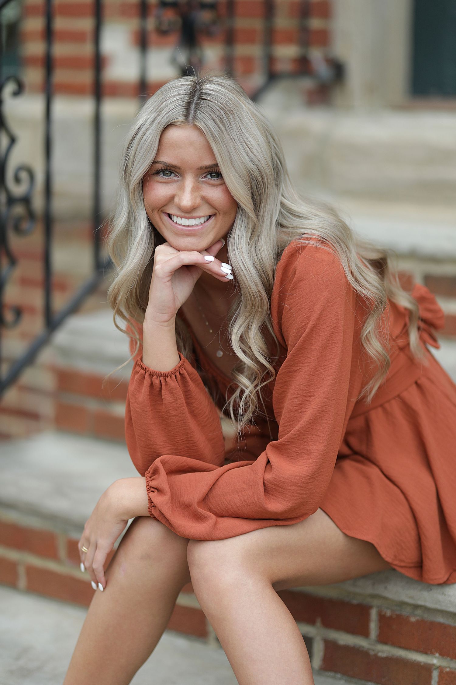 Young woman in rust-colored sweater dress sitting on brick steps with long wavy platinum blonde hair and bright smile.