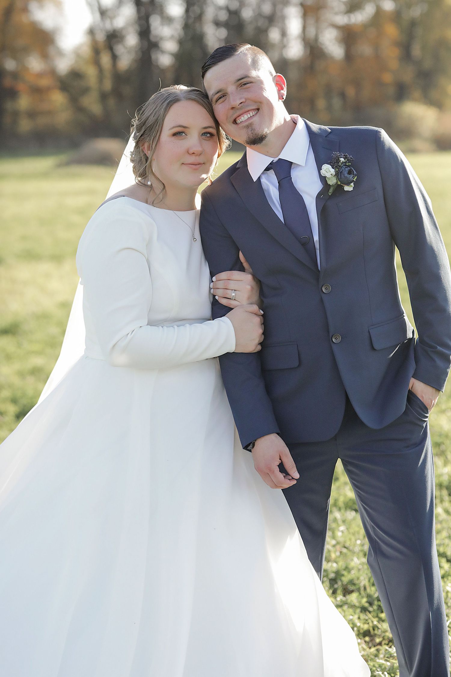 A wedding couple poses together outdoors during autumn, wearing classic formal attire.