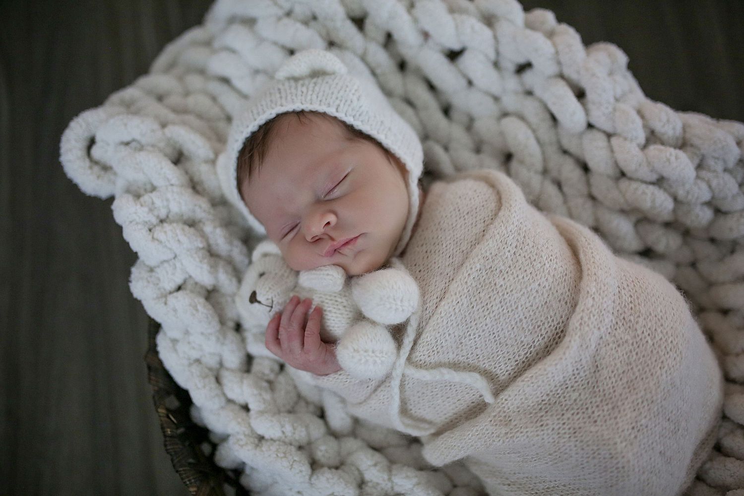Newborn baby wrapped in cozy cream knitted blanket sleeps peacefully on textured white backdrop.