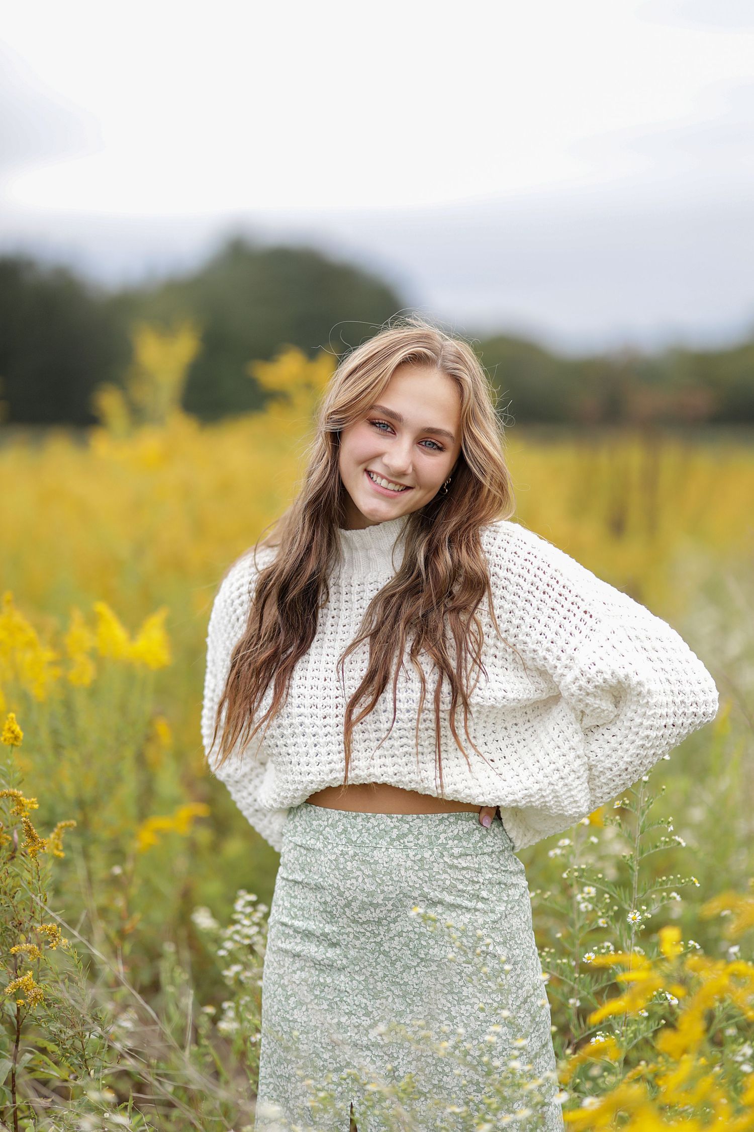 A person in a white knit sweater poses joyfully in a field of blooming yellow wildflowers on a cloudy day.