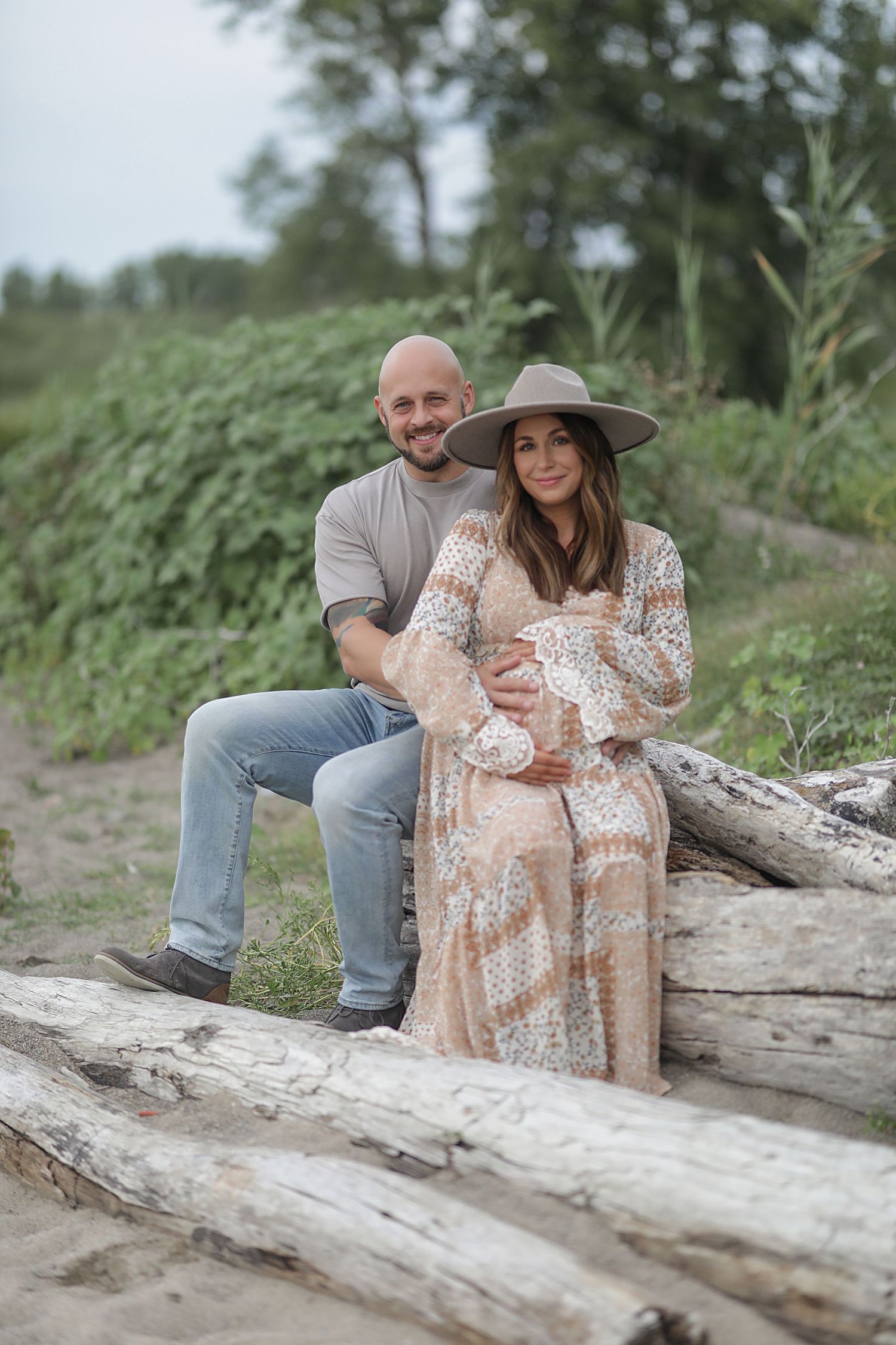 Couple sits together on driftwood log at beach, wearing casual attire with woman in flowy dress and wide-brim hat.
