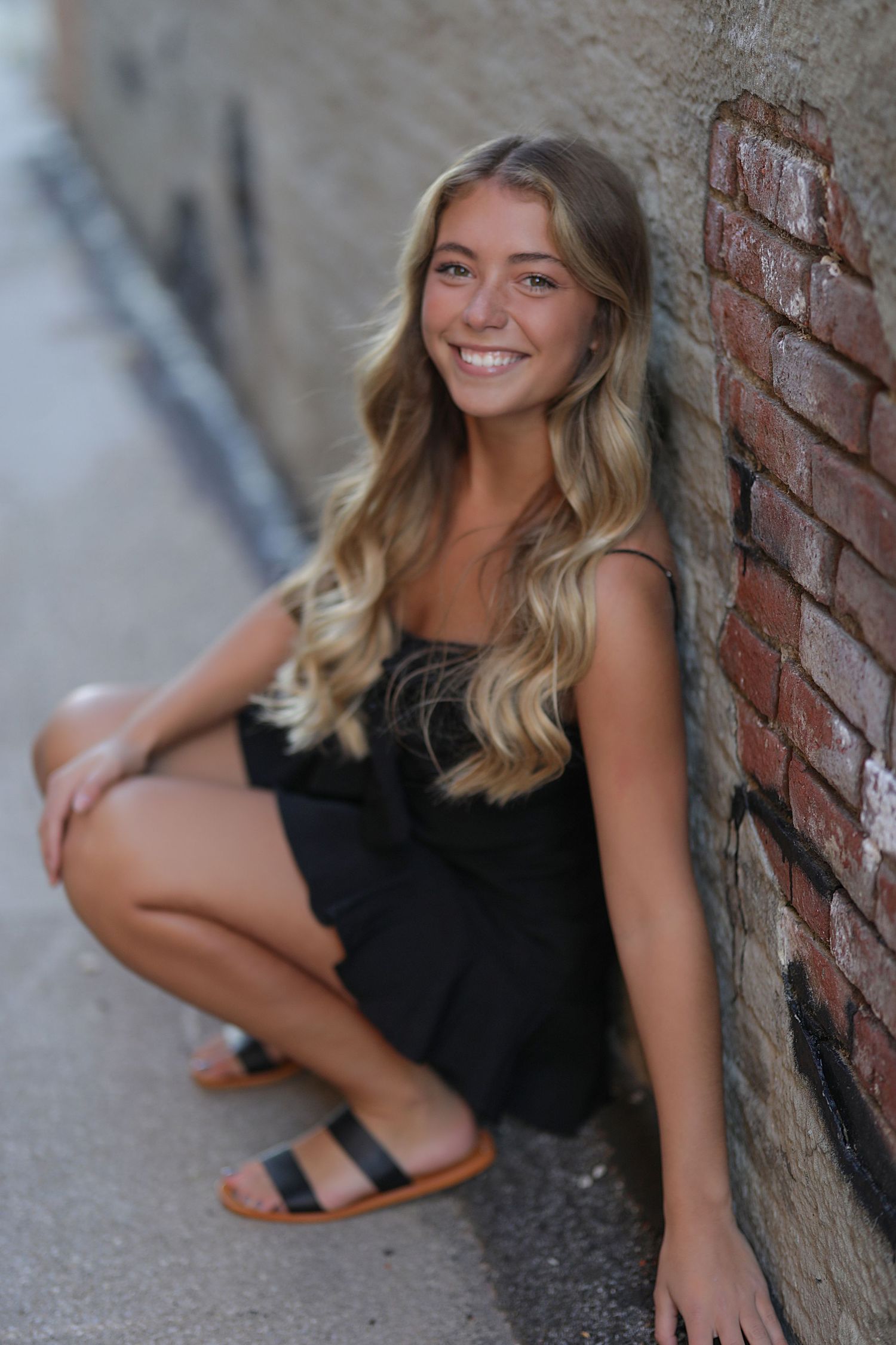Young woman with long blonde wavy hair wearing black dress crouches by brick wall outdoors smiling at camera.