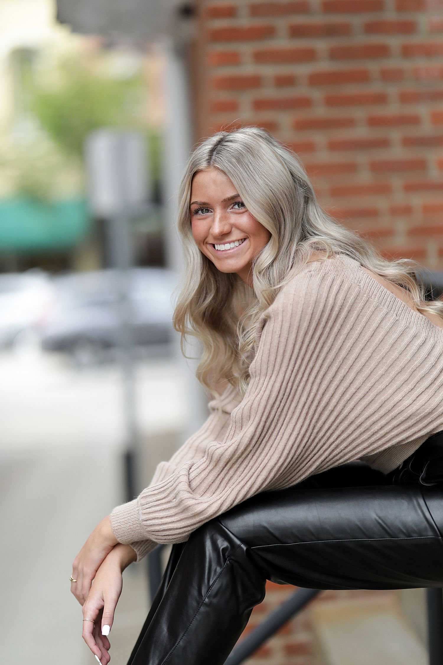 Fashion model wearing a cozy beige ribbed sweater and leather pants poses casually against a brick wall background.