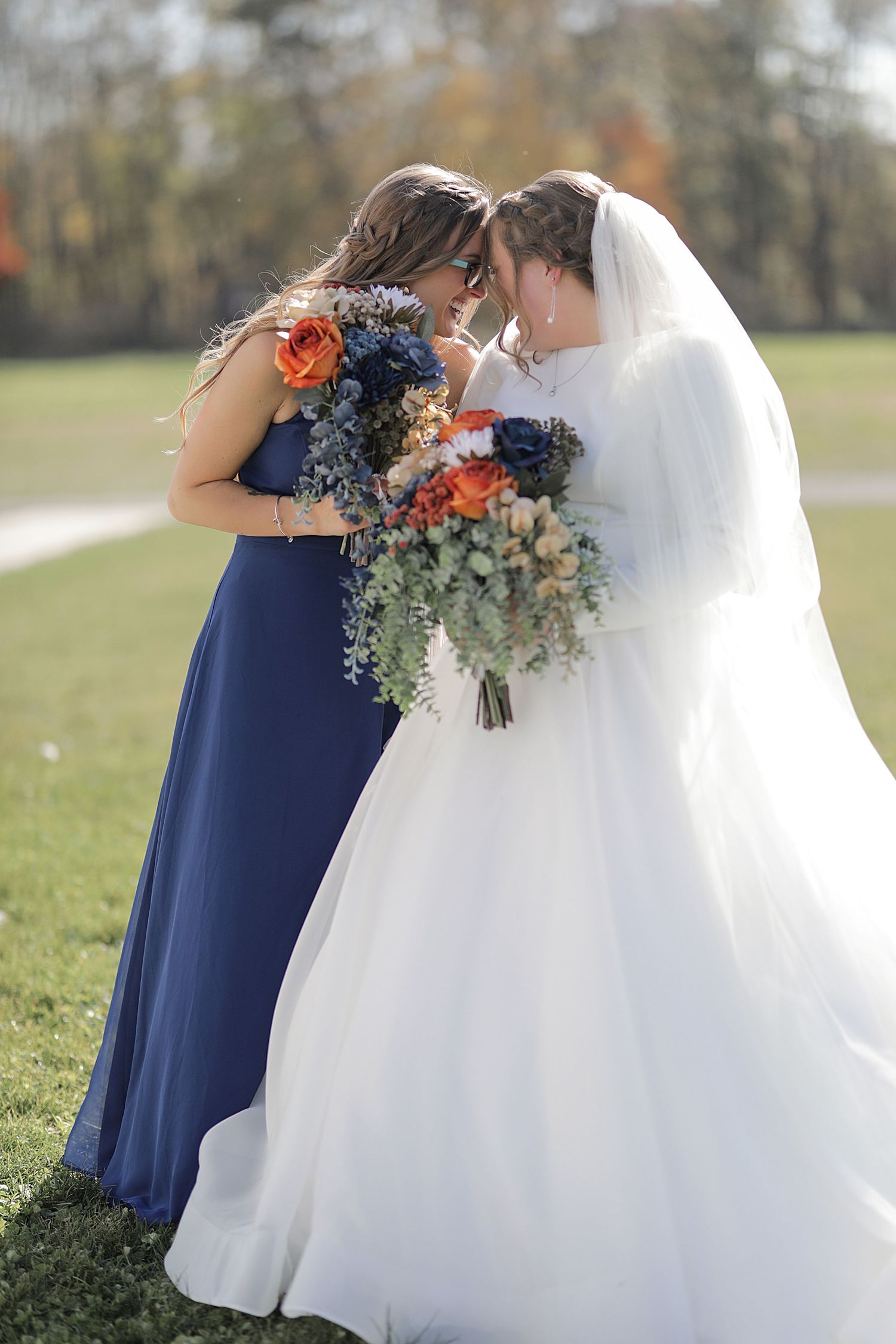 Two wedding attendees share a tender moment outdoors while holding beautiful autumn-colored bouquets.
