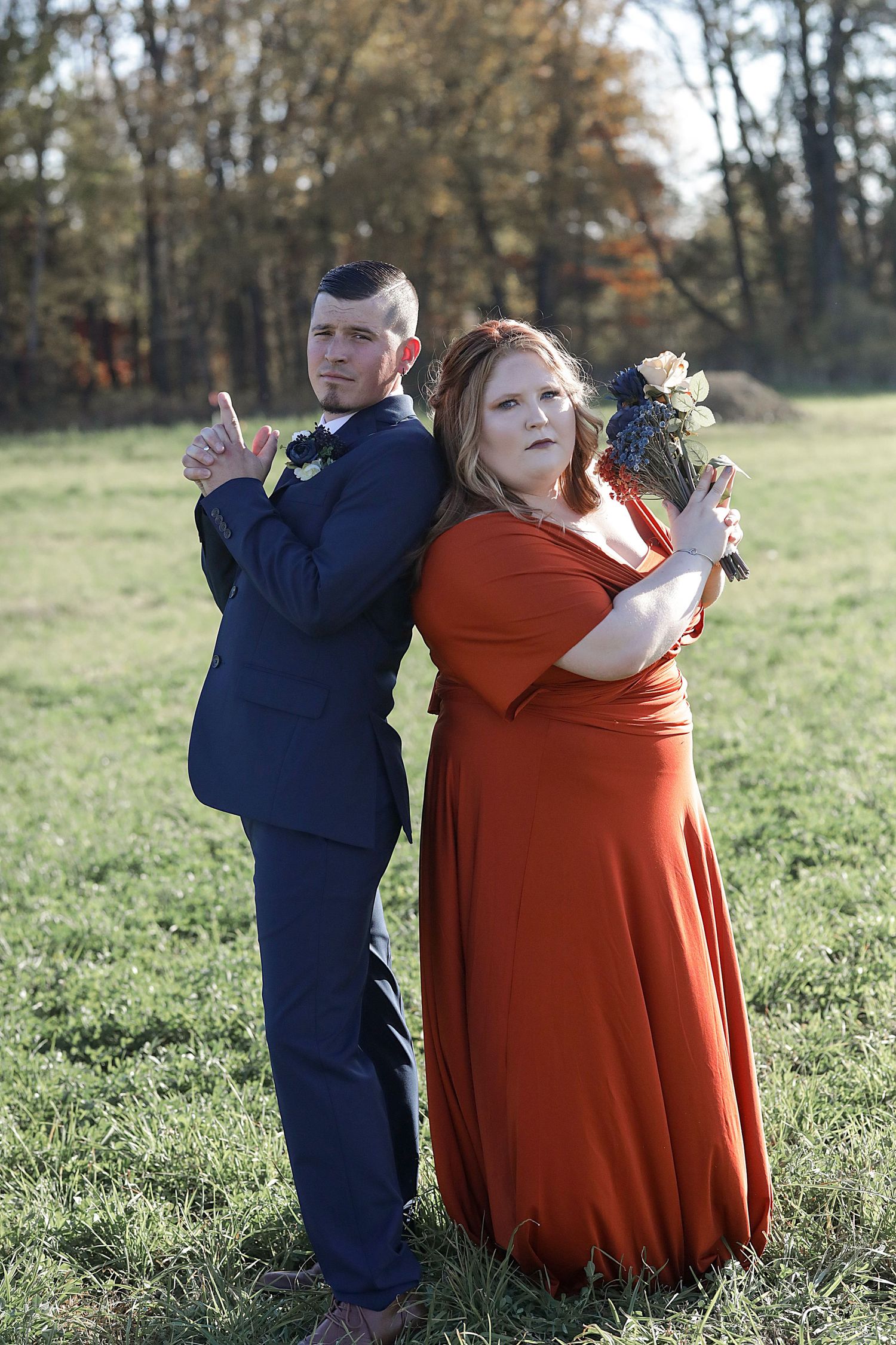 A playful wedding photo with couple posing back-to-back in a field, showing navy suit and burnt orange dress.