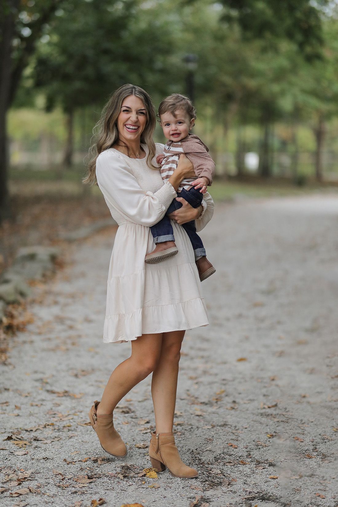 Parent wearing white dress smiles while holding laughing toddler on tree-lined fall path.