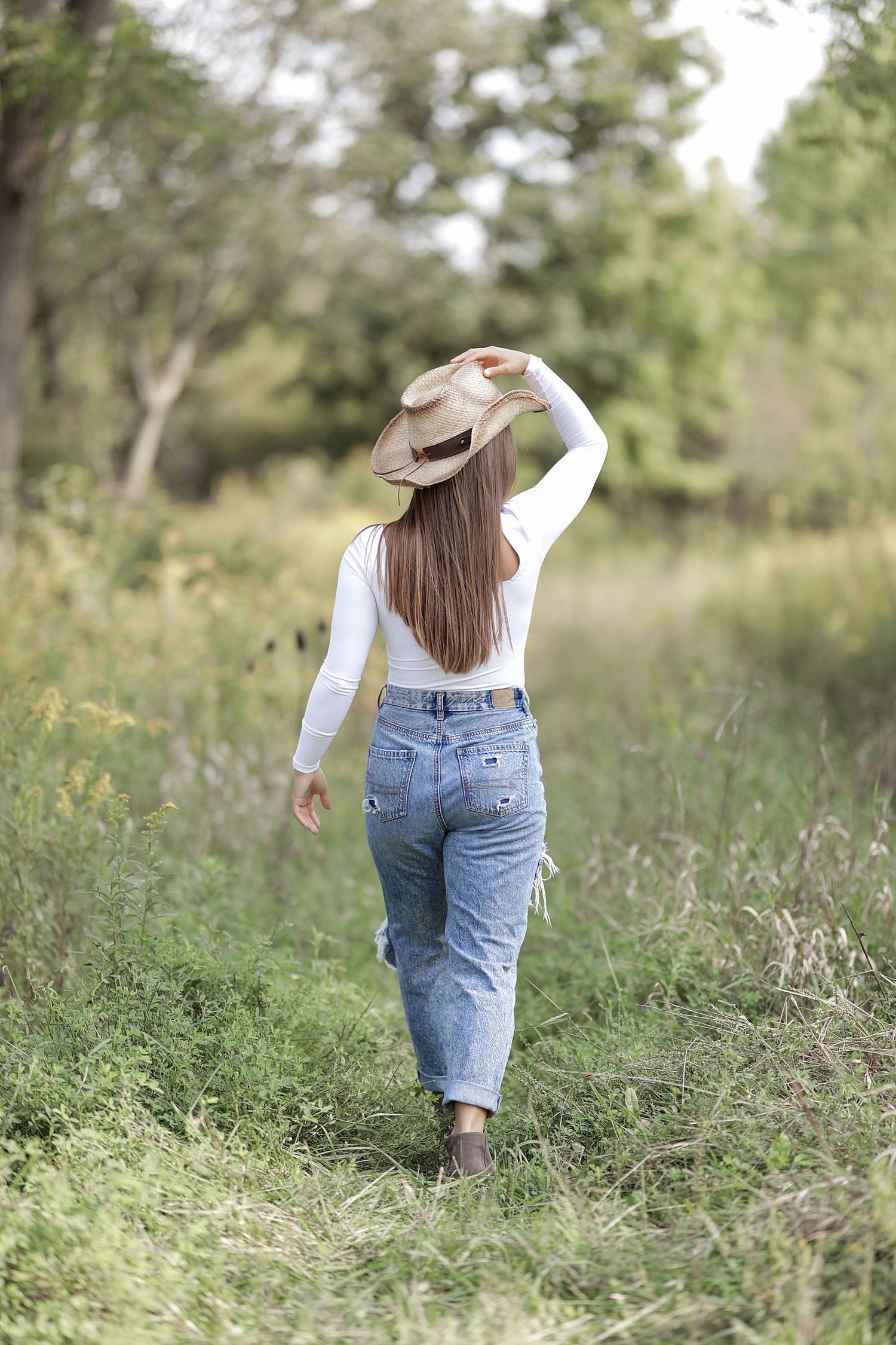 Woman in white top and blue jeans walking through a meadow while holding a brown hat on a sunny summer day.