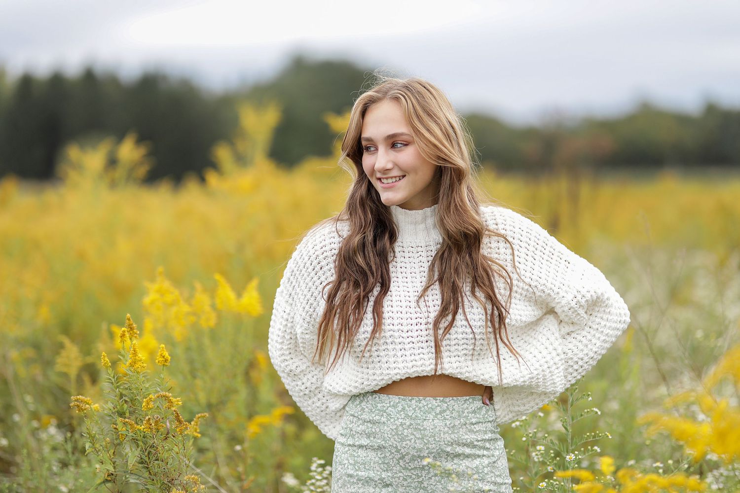 A person in a white knit sweater poses in a field of blooming yellow wildflowers on an overcast day.