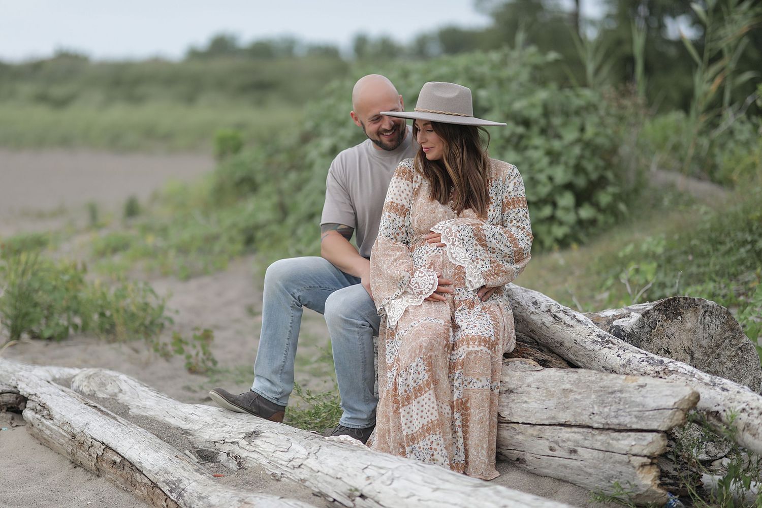 A couple sits together on driftwood along a coastal trail during a maternity photo session in nature.