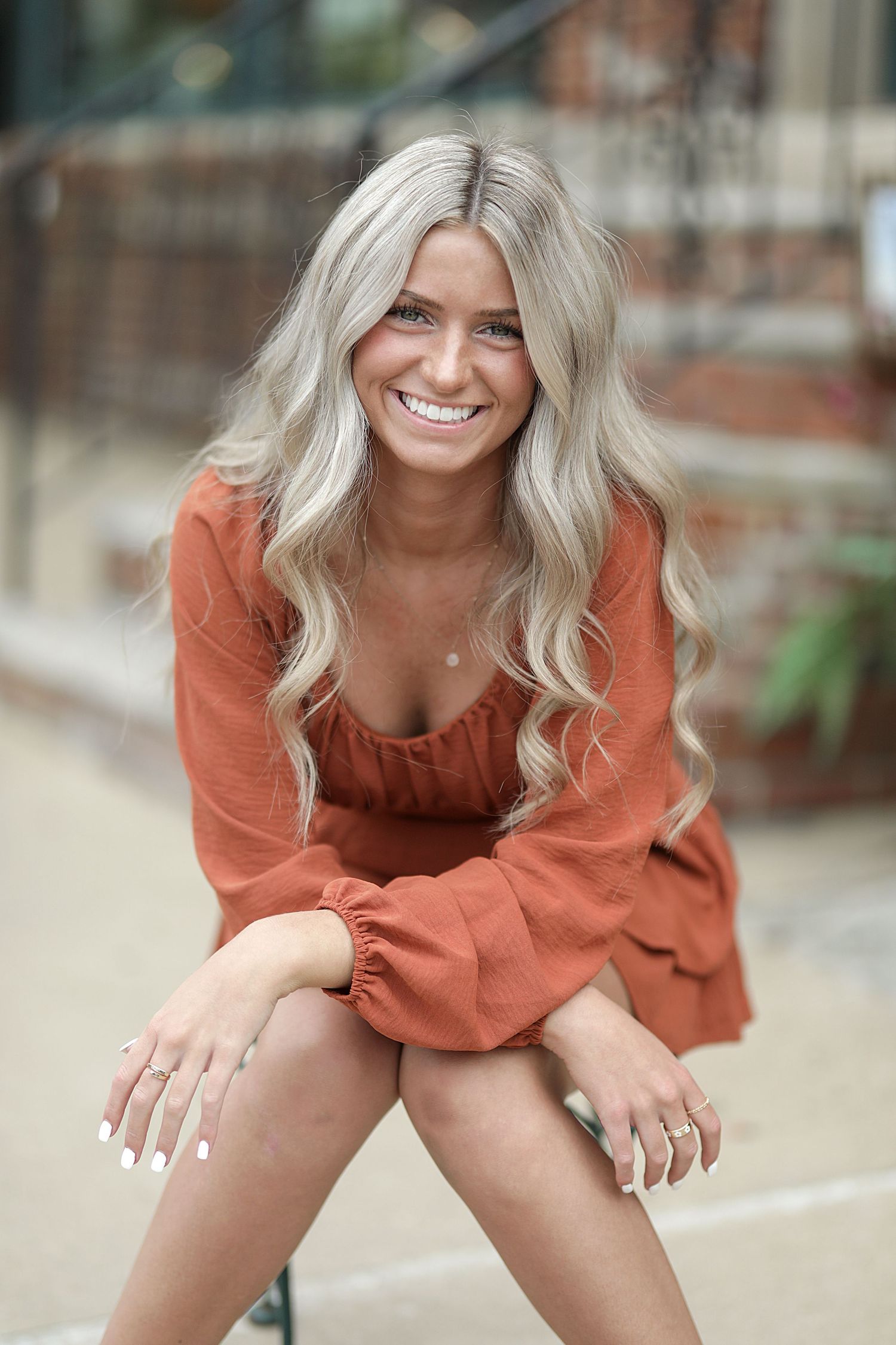 Outdoor portrait of person with platinum blonde wavy hair wearing a rust-colored dress with billowy sleeves.