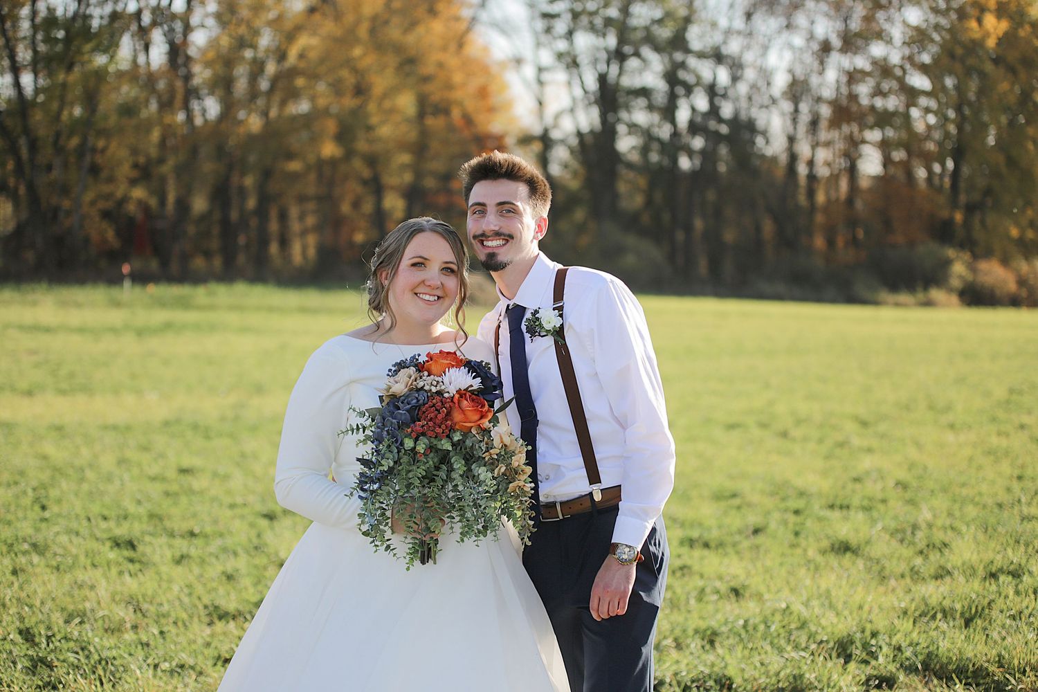 Newlyweds walking through sunny field with rustic fall foliage in background.