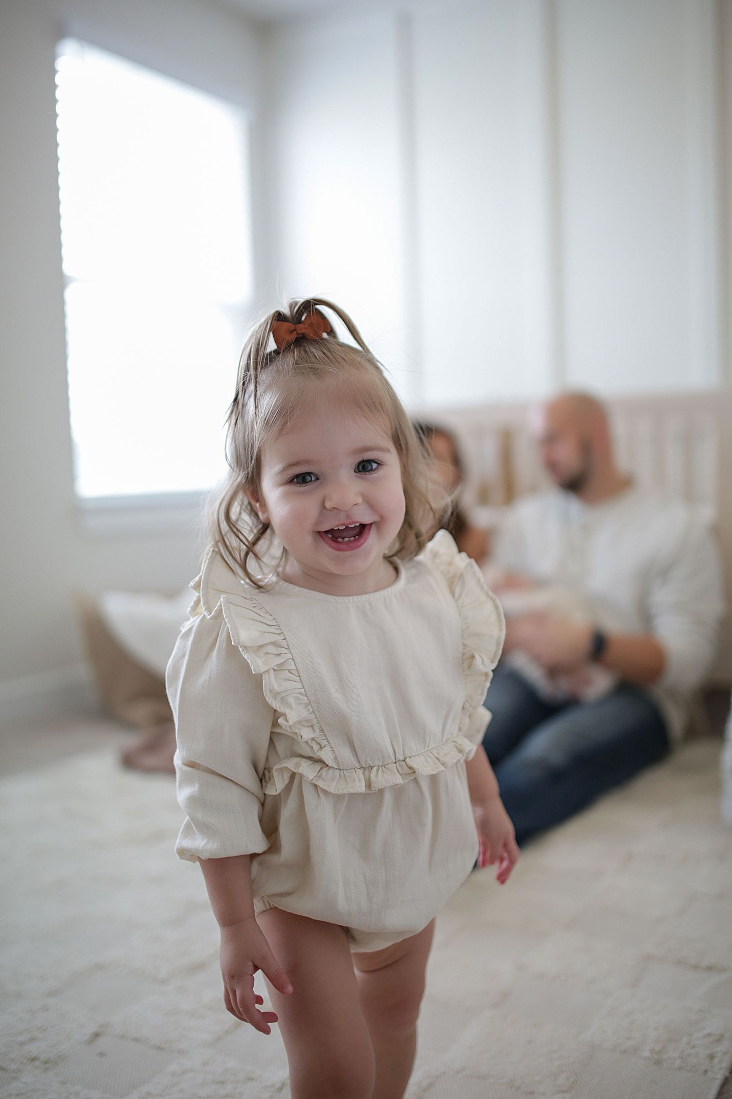A joyful toddler wearing a cream-colored ruffled top smiles brightly in a bright, minimalist interior setting.
