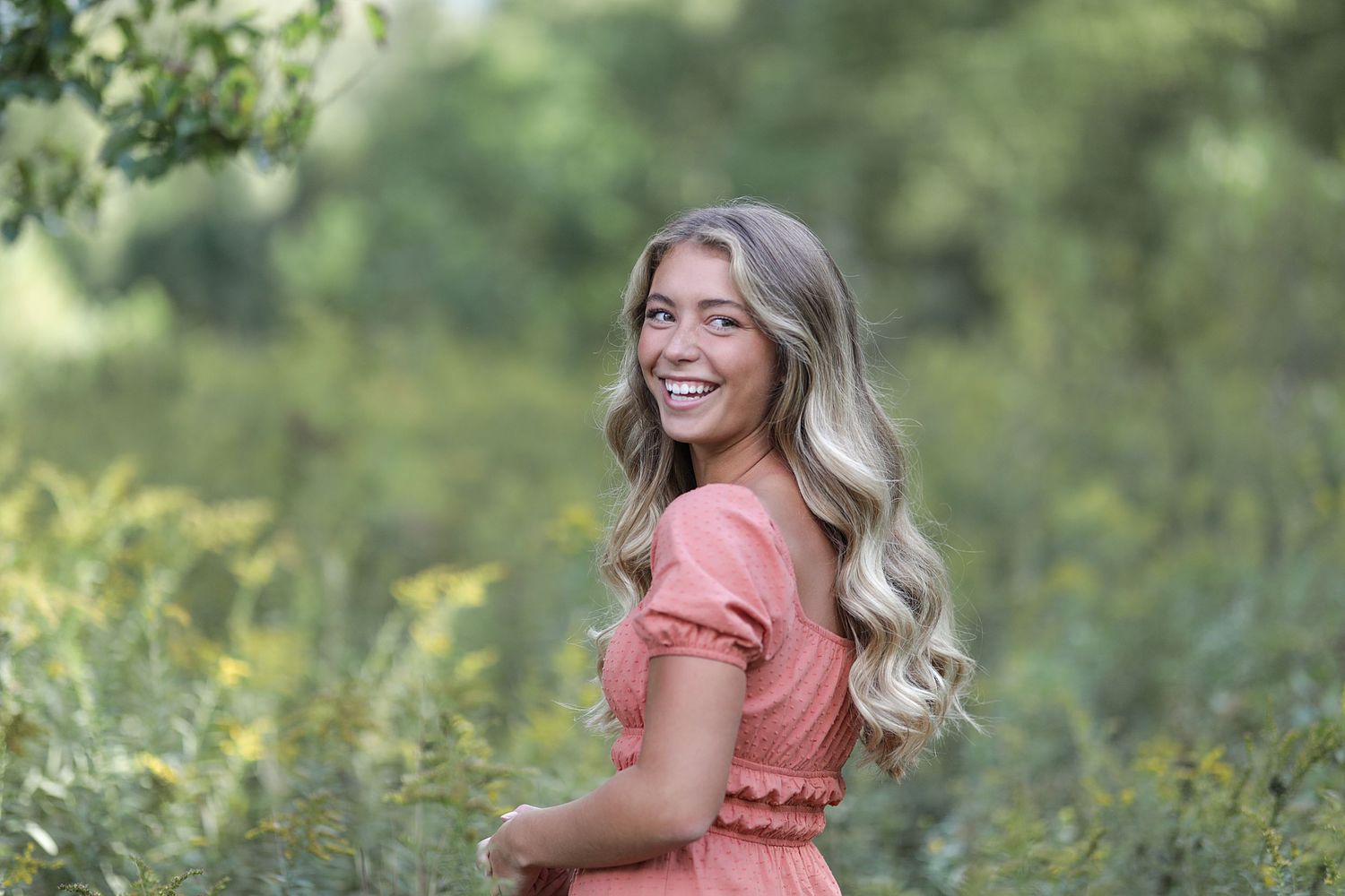 A woman in a coral pink dress smiles outdoors surrounded by greenery in a natural setting.