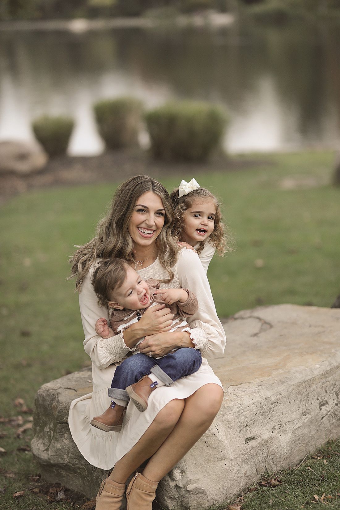 Mother with two children sits on rock by lake during family portrait session.