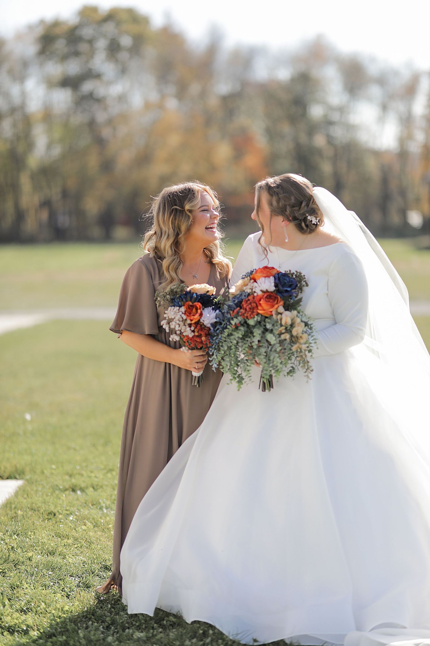 Two wedding attendees share joyful laughter while holding vibrant fall bouquets in an outdoor setting.