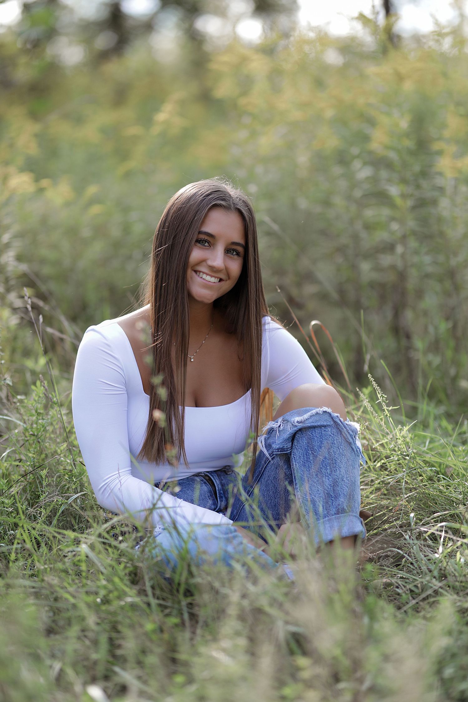 A young woman sits in long grass wearing a white long-sleeve top and blue jeans on a sunny afternoon.