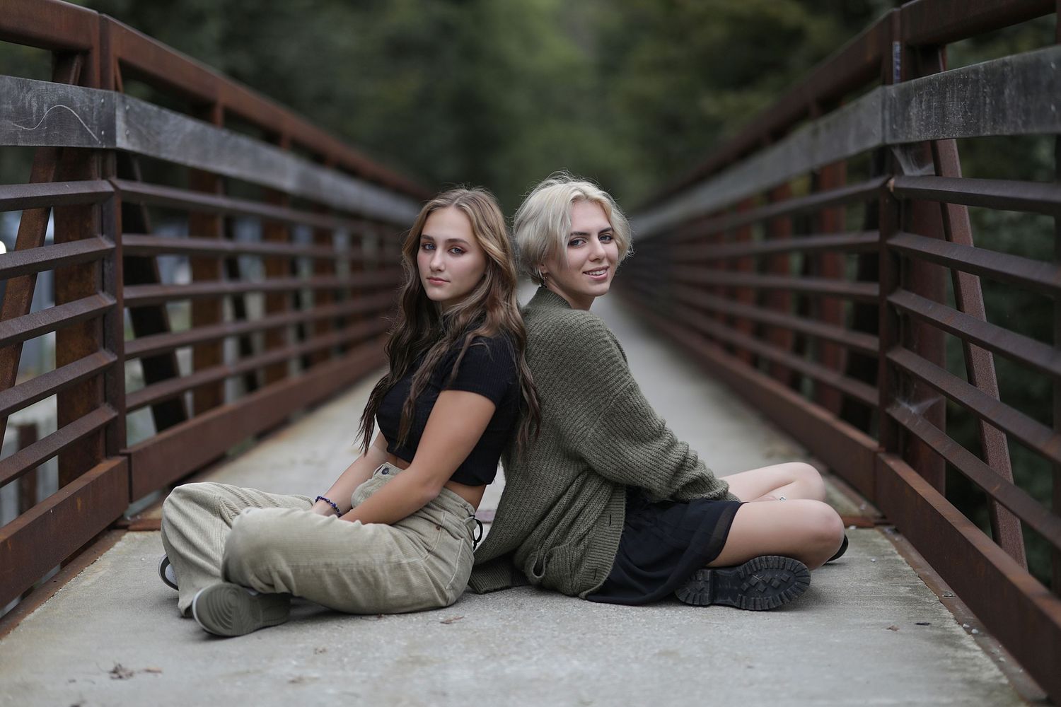 Two friends in casual outfits lie together on a wooden bridge while taking photos in nature.