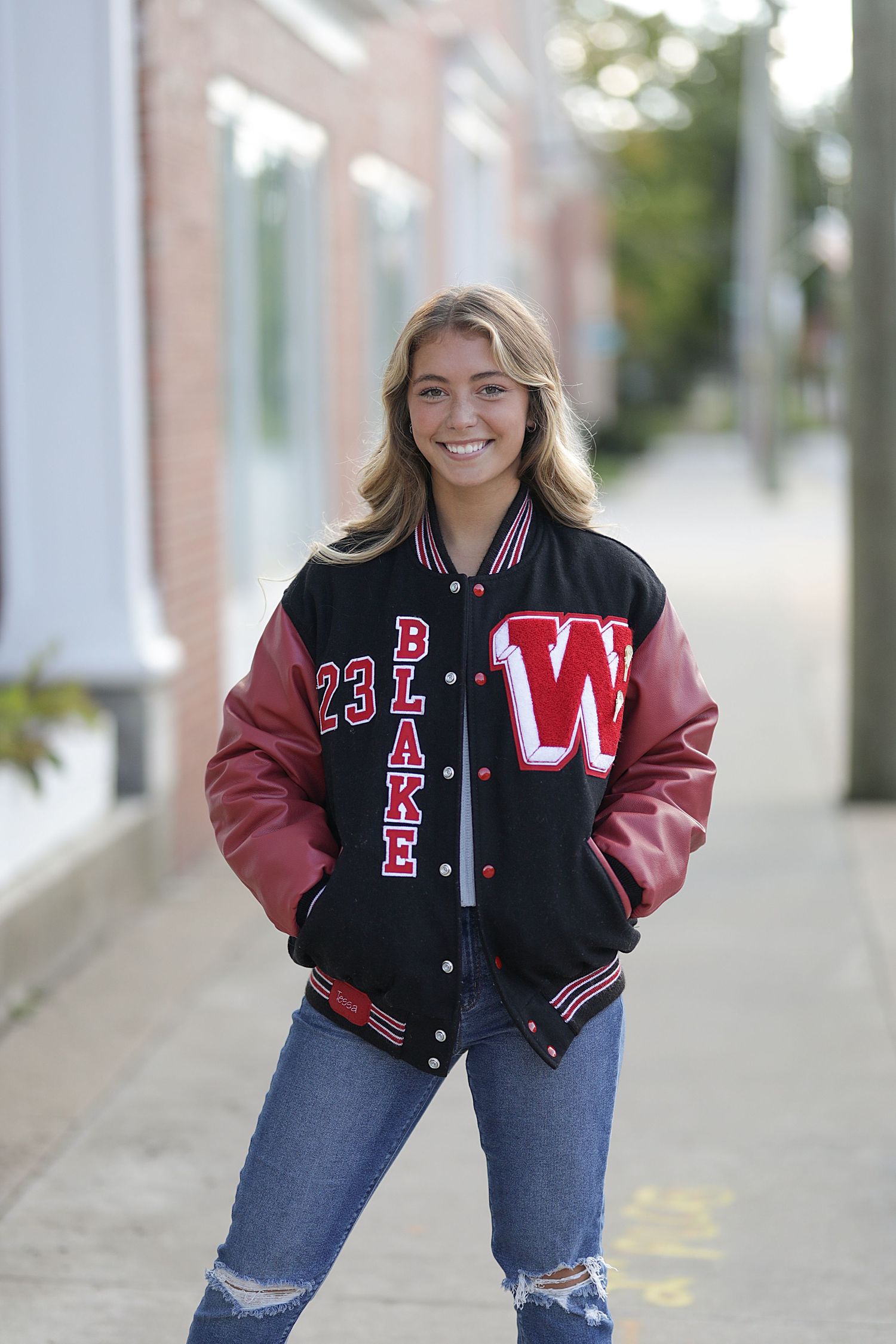 Person wearing a black and burgundy varsity letterman jacket with blue ripped jeans in an outdoor setting with columns.